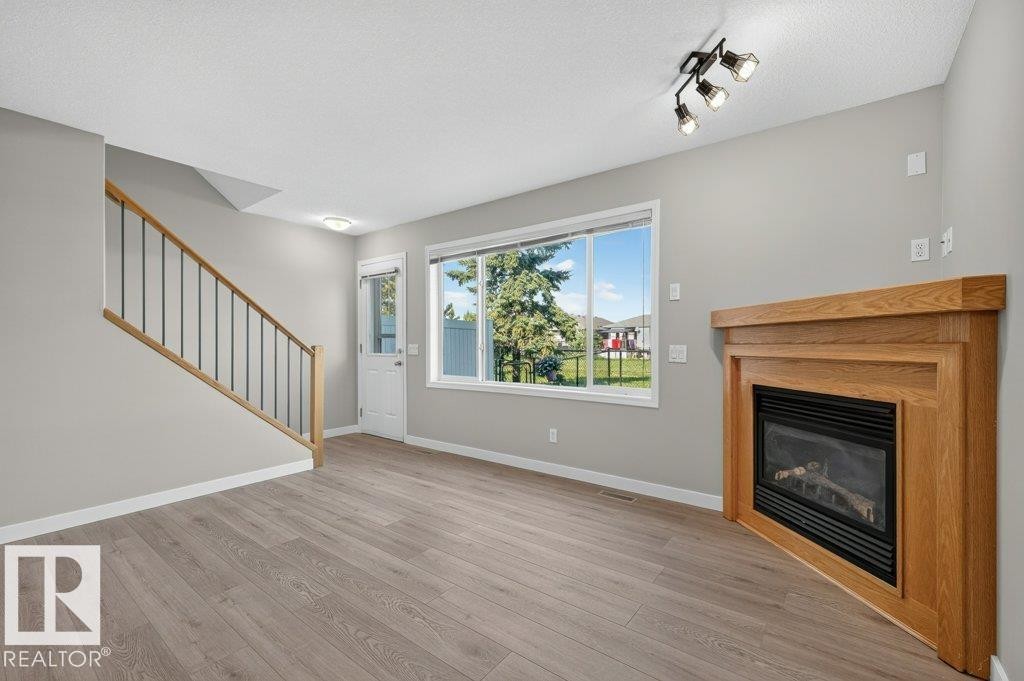 Wood-finish flooring, staircase with wood handrail and black spindles, built-in fireplace with wood mantel, large window, and track lighting fixture - #26 2816 34 Av Nw, Edmonton, AB