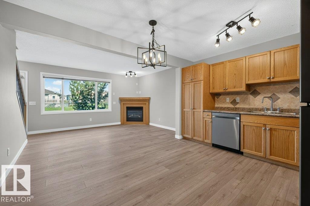 Wood-finish flooring throughout, featuring a corner fireplace with a wooden mantel, a large window, a kitchen area with wood cabinetry, and stainless steel appliances - #26 2816 34 Av Nw, Edmonton, AB