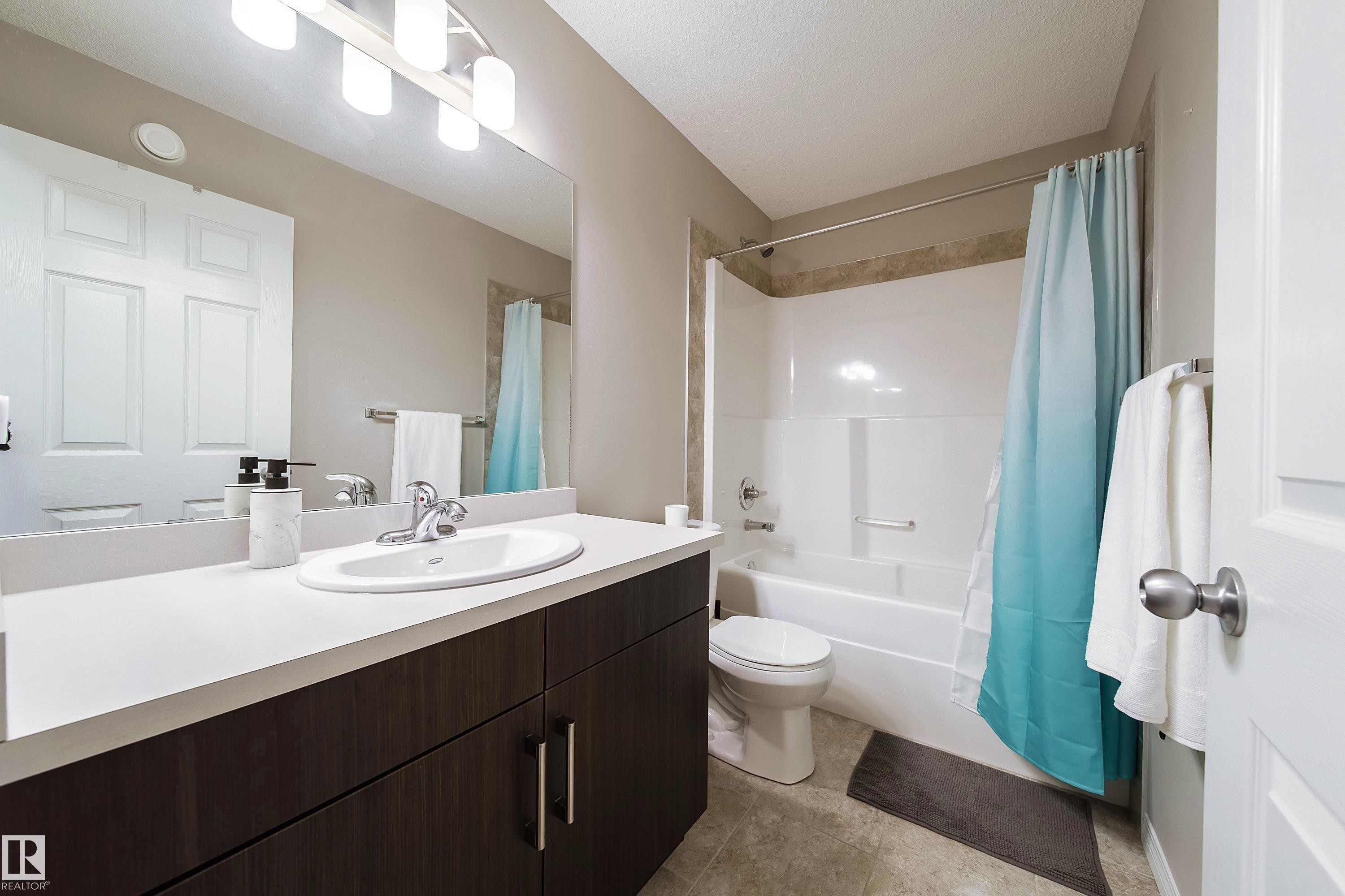Dark wood-finish cabinetry with silver hardware, white countertop with integral sink, large vanity mirror, four-bulb vanity lighting fixture, and a combination tub and shower - 3211 Cherry Crescent Sw, Edmonton, AB