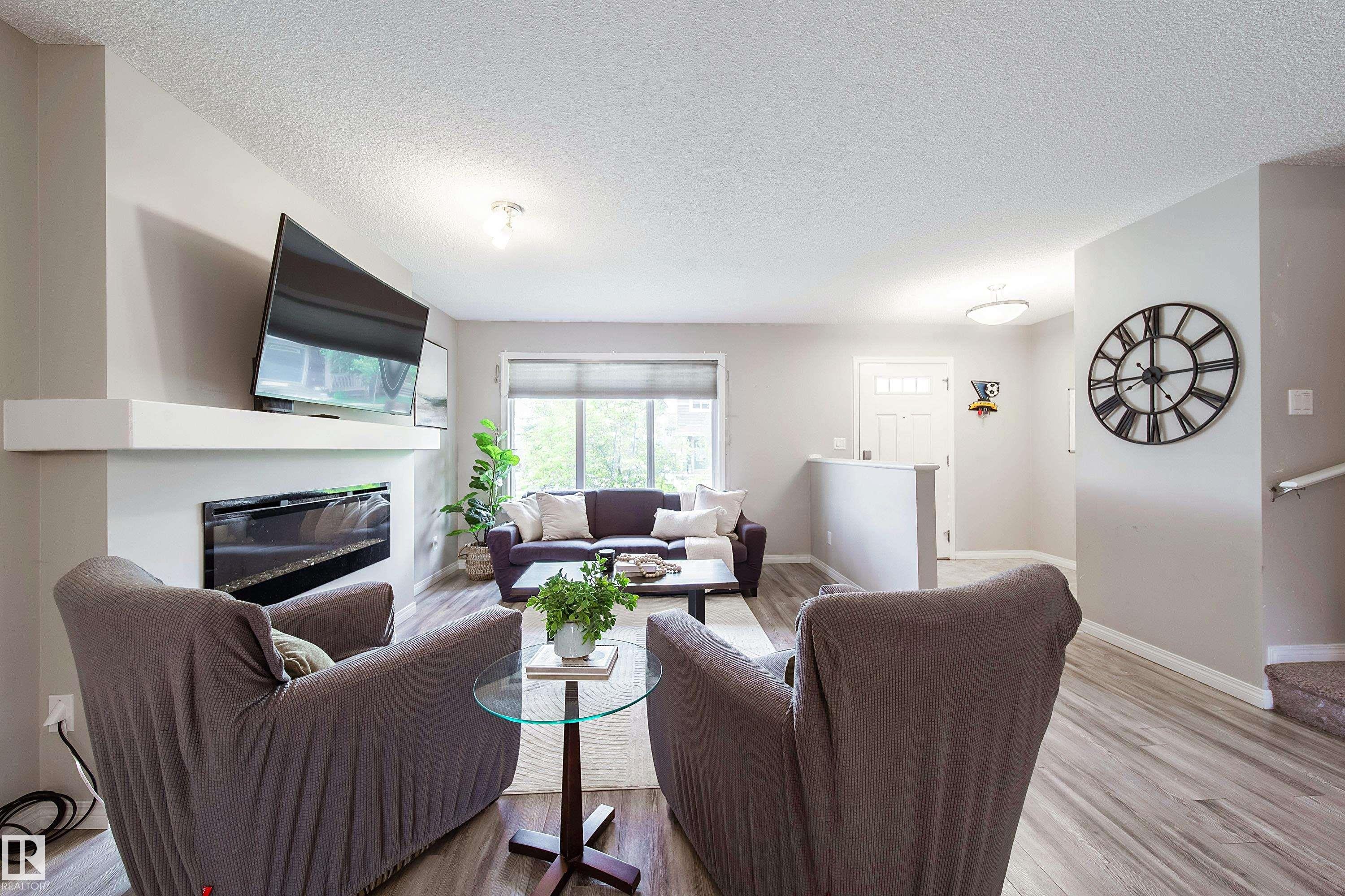 Modern open-concept living space featuring a wood-finish plank floor, gas fireplace with built-in cabinetry, wall-mounted flat-screen television, and abundant natural light from large picture windows - 3211 Cherry Crescent Sw, Edmonton, AB
