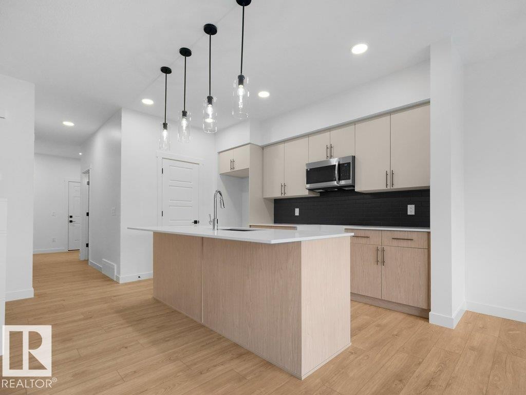 Modern kitchen featuring a central island with a sink and white countertop, light wood-finish cabinetry, a dark tile backsplash, and stainless steel appliances - 912 17 Ave Nw, Edmonton, AB