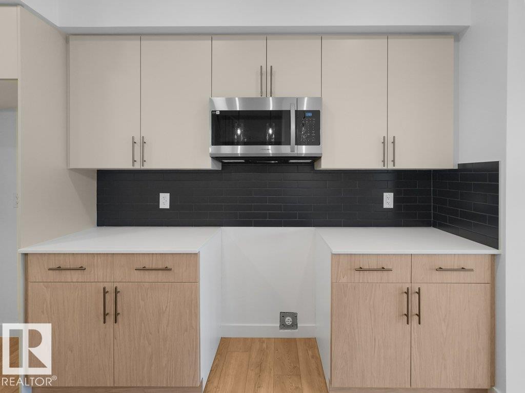 Sleek kitchen featuring light-toned upper cabinetry, stainless steel microwave, black subway tile backsplash, white countertops, and wood-finish lower cabinetry - 912 17 Ave Nw, Edmonton, AB