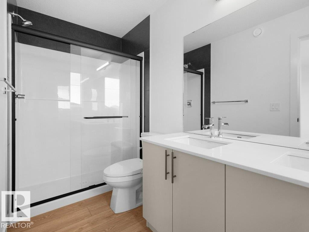 Dual basin vanity with light-toned cabinetry and chrome fixtures, frameless wall-mounted mirror, wood-finish flooring, and a glass-enclosed shower with black trim - 912 17 Ave Nw, Edmonton, AB