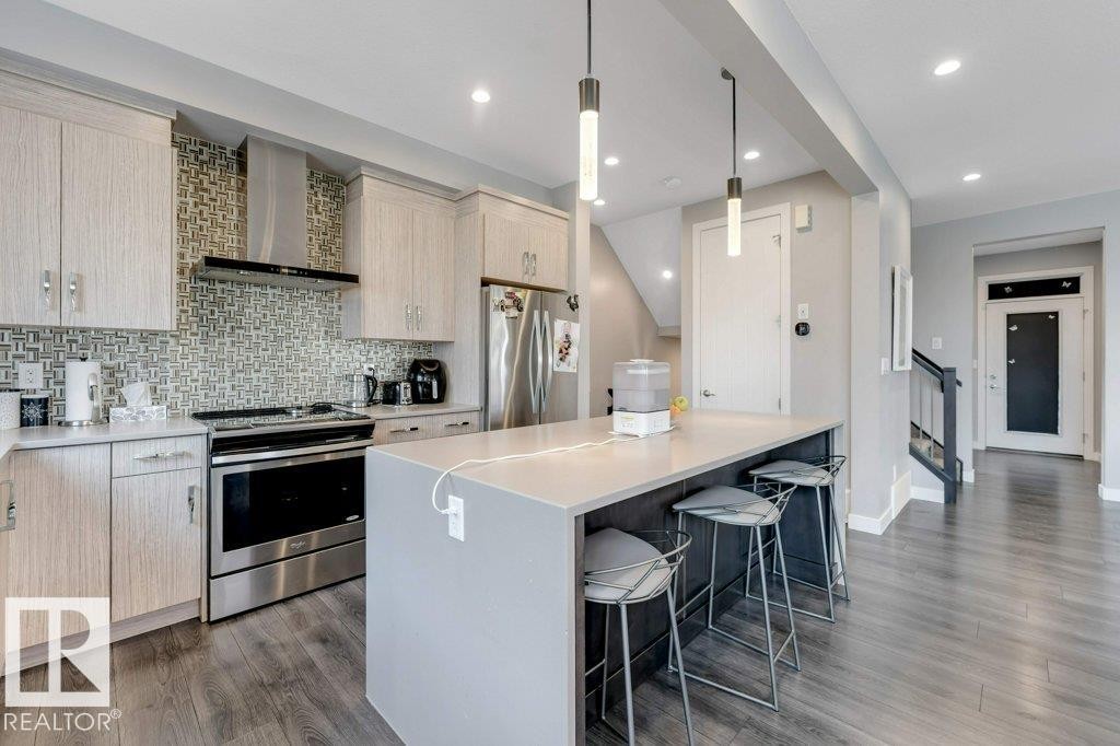 Kitchen featuring light wood-finish cabinetry, stainless steel appliances, a tiled backsplash, a kitchen island with a light countertop, and recessed lighting - #45 16903 68 St Nw, Edmonton, AB