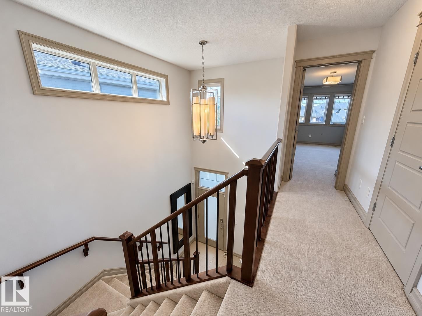 Carpeted staircase with dark wood-finish handrails and balusters, transom windows with light wood-finish trim, and a decorative pendant light fixture - 885 Windermere Wynd Nw, Edmonton, AB