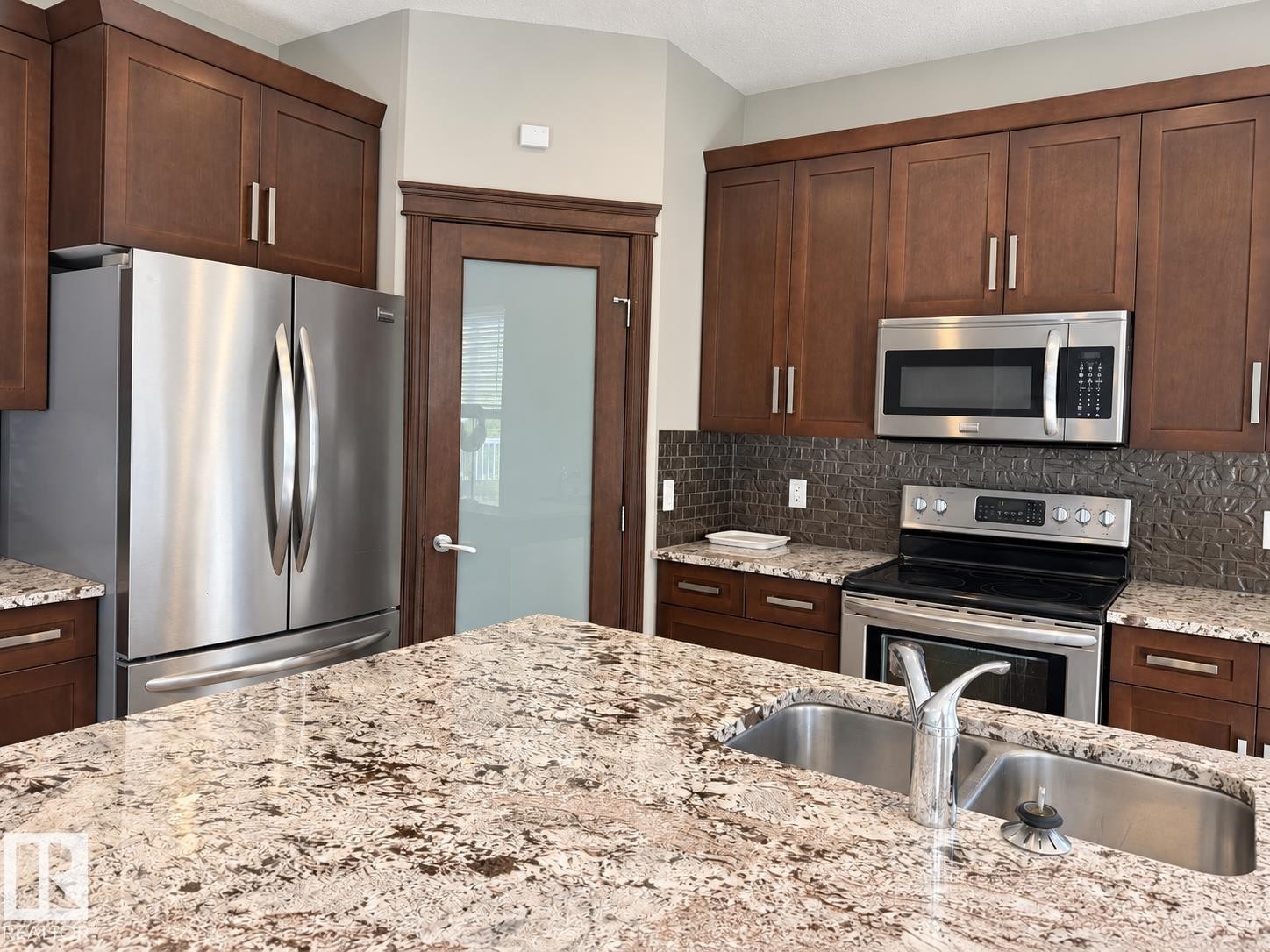 Kitchen featuring wood cabinetry, stainless steel appliances, speckled stone countertops, a double basin sink with a high-arc faucet, and textured backsplash - 885 Windermere Wynd Nw, Edmonton, AB