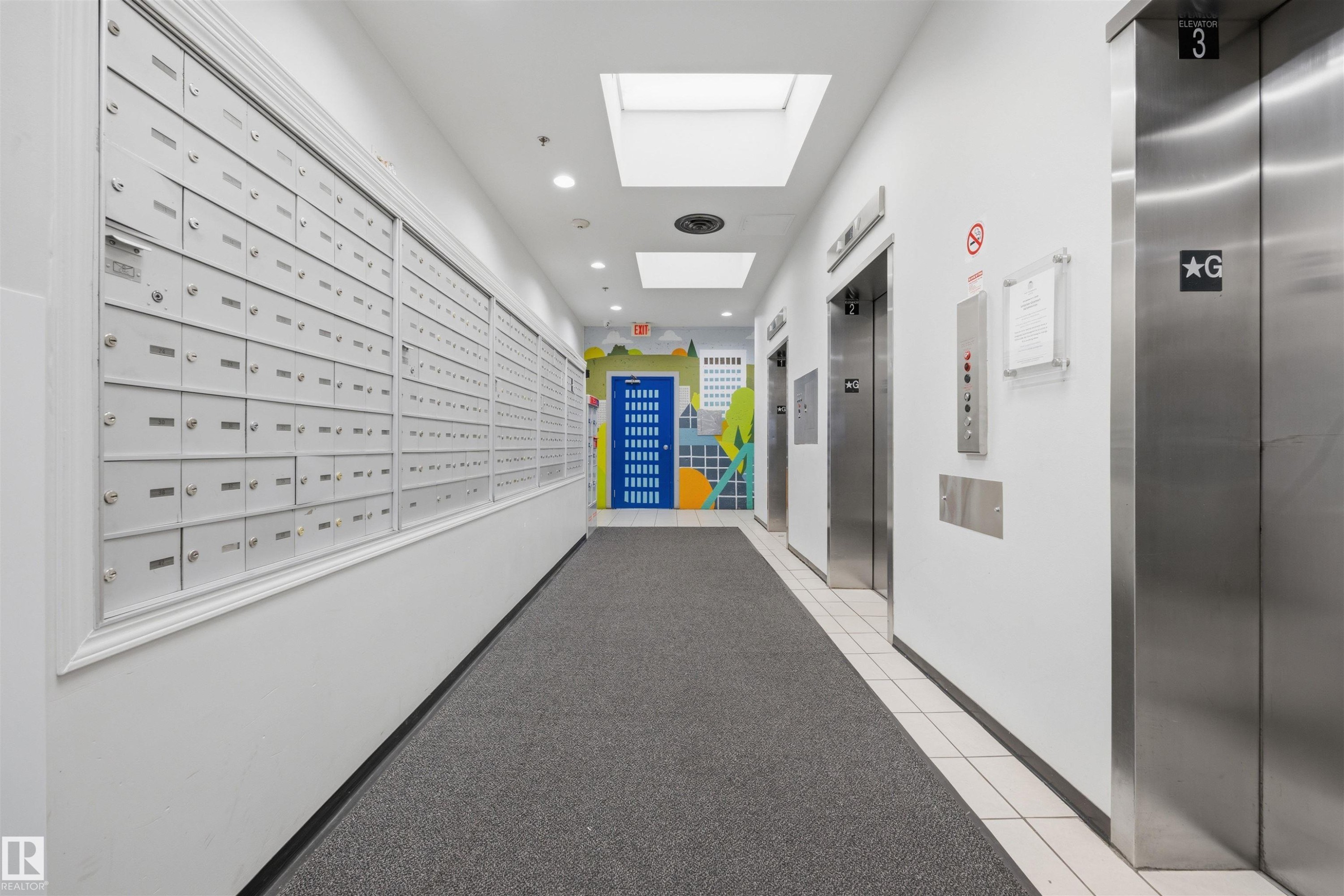 Modern building corridor featuring wall-mounted mailboxes with individual compartments, polished stainless steel elevator doors, recessed ceiling lighting, neutral wall finishes, and gray tile flooring throughout - #1313 10024 Jasper Av Nw, Edmonton, AB
