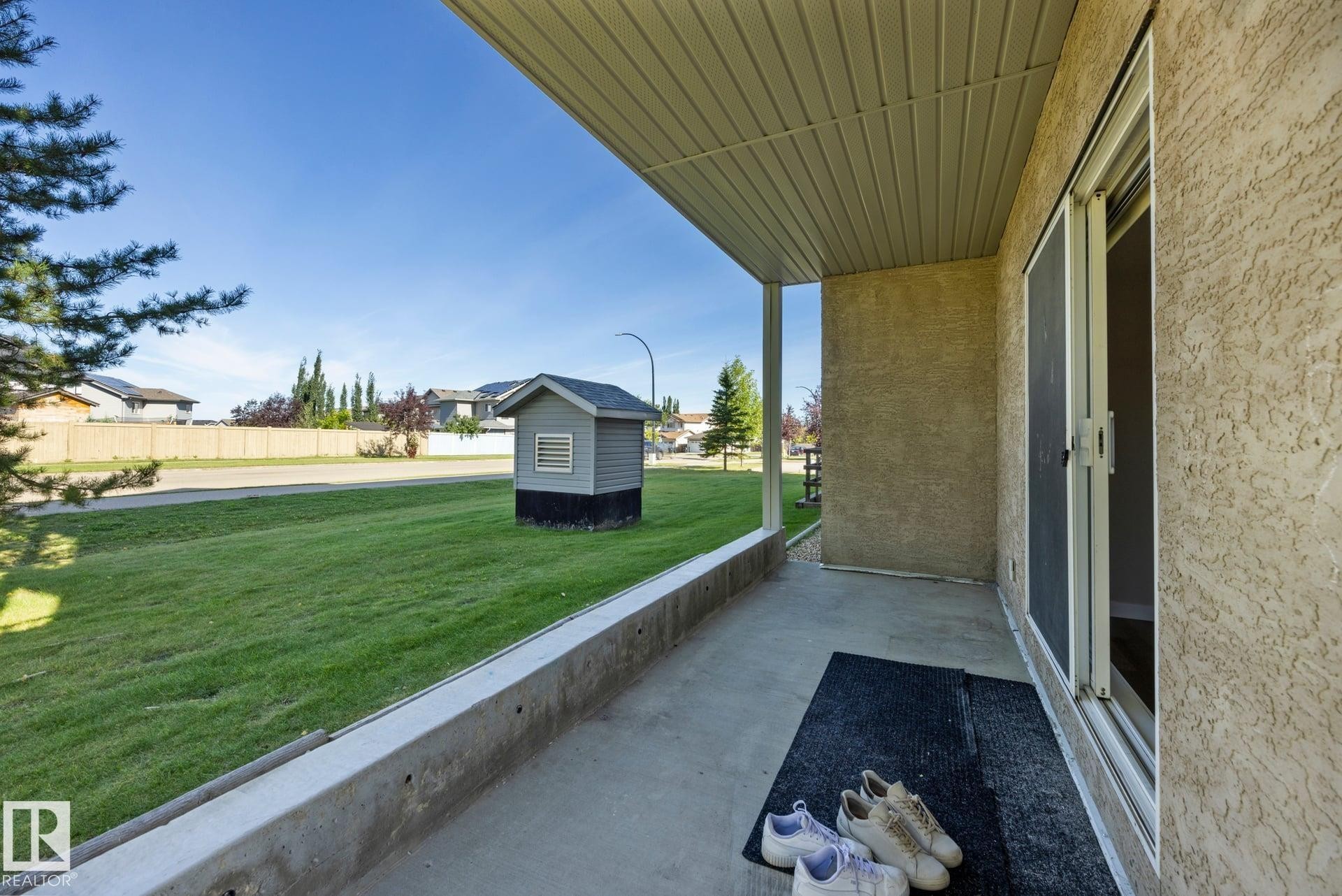 Covered concrete patio with sliding glass door access, stucco exterior, and a manicured lawn - #122 240 Spruce Ridge Rd, Spruce Grove, AB