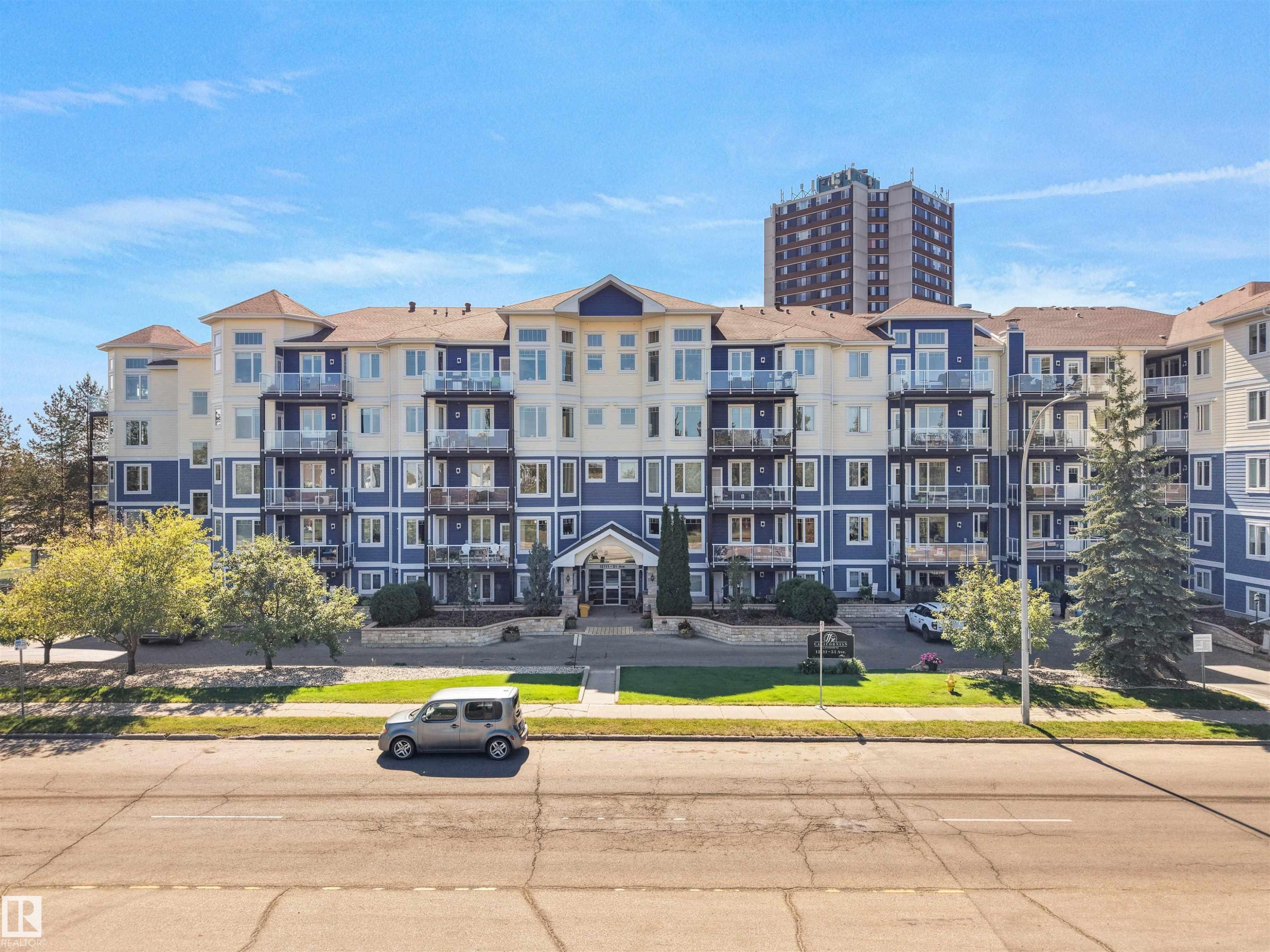Multi-story building featuring blue and cream-colored siding, multiple glass-paneled balconies, and a paved street - #206 12111 51 Av Nw, Edmonton, AB