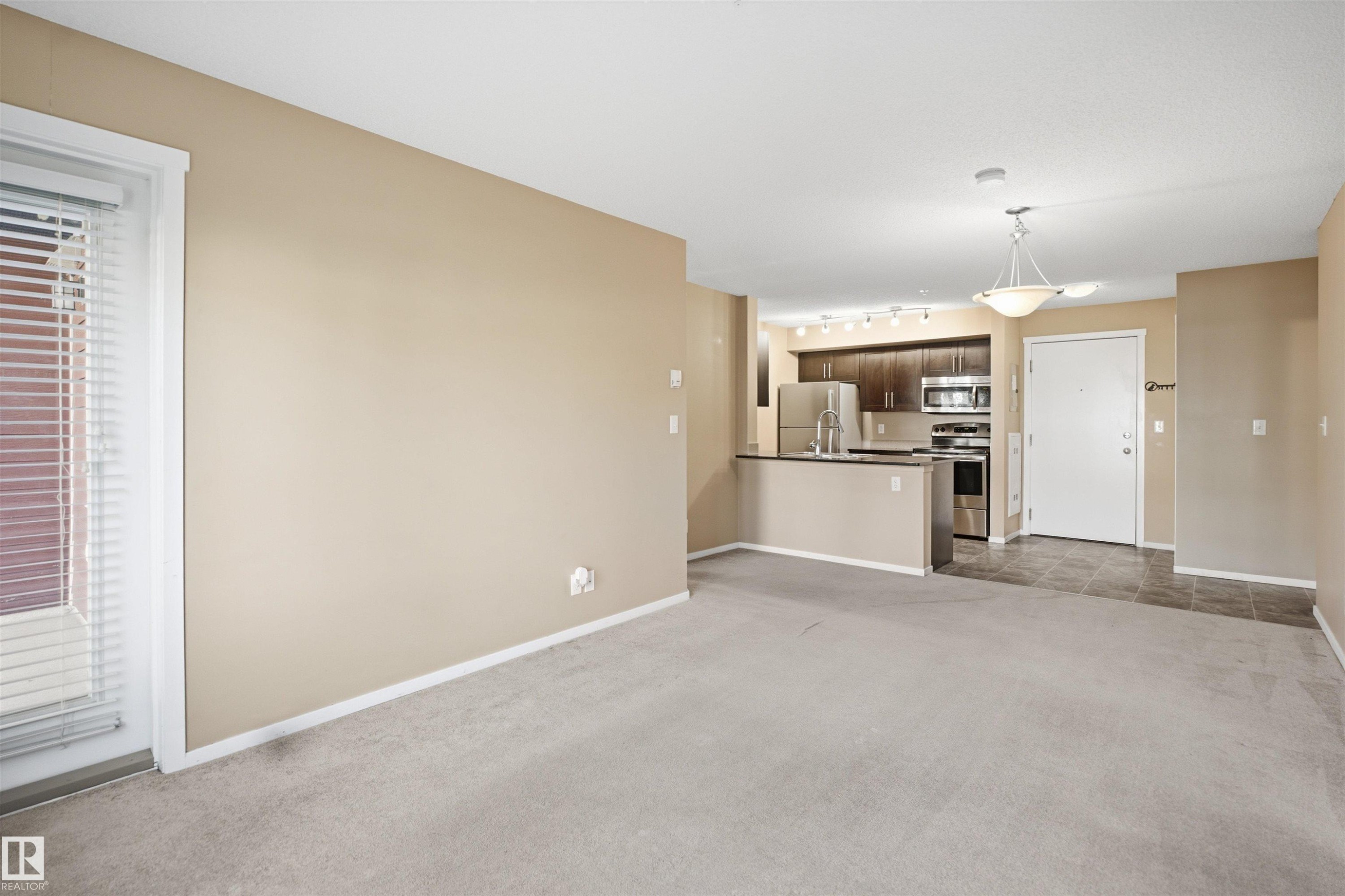 Open-concept living area with neutral carpeting and a kitchen featuring dark wood-finish cabinetry, stainless steel appliances, and a breakfast bar - #225 1510 Watt Dr Sw, Edmonton, AB