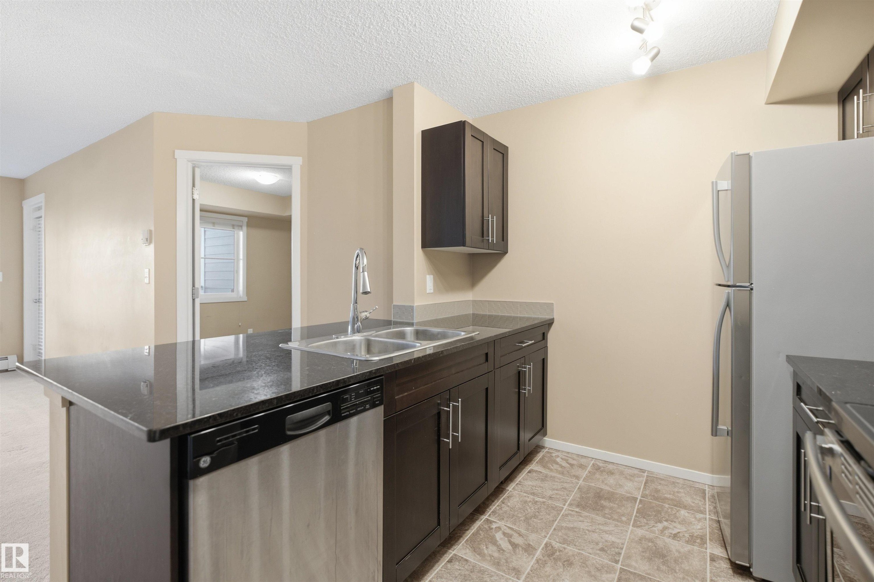 Kitchen featuring dark wood-finish cabinetry, stainless steel appliances, a dual basin sink, and tile flooring - #225 1510 Watt Dr Sw, Edmonton, AB