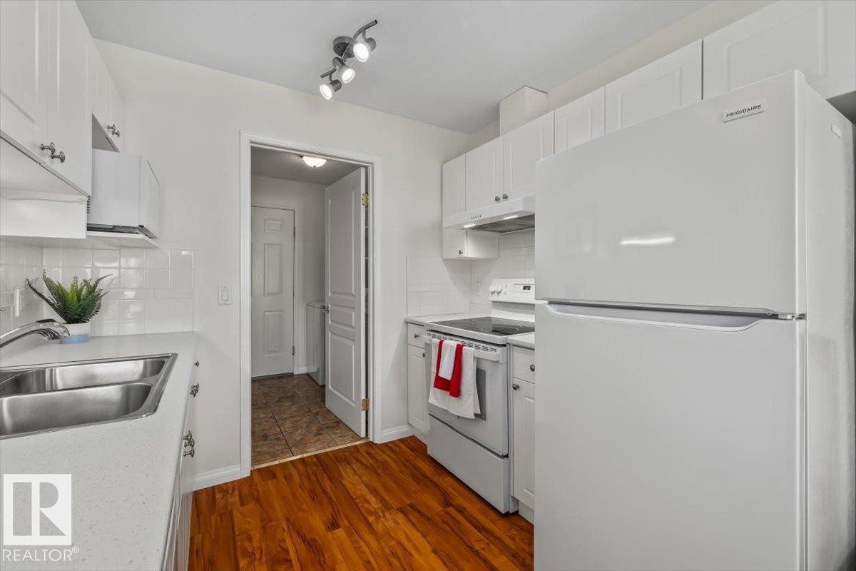 Kitchen featuring white cabinetry, a double basin stainless steel sink, white appliances, track lighting, and wood-finish flooring - #92 13320 124 St Nw, Edmonton, AB