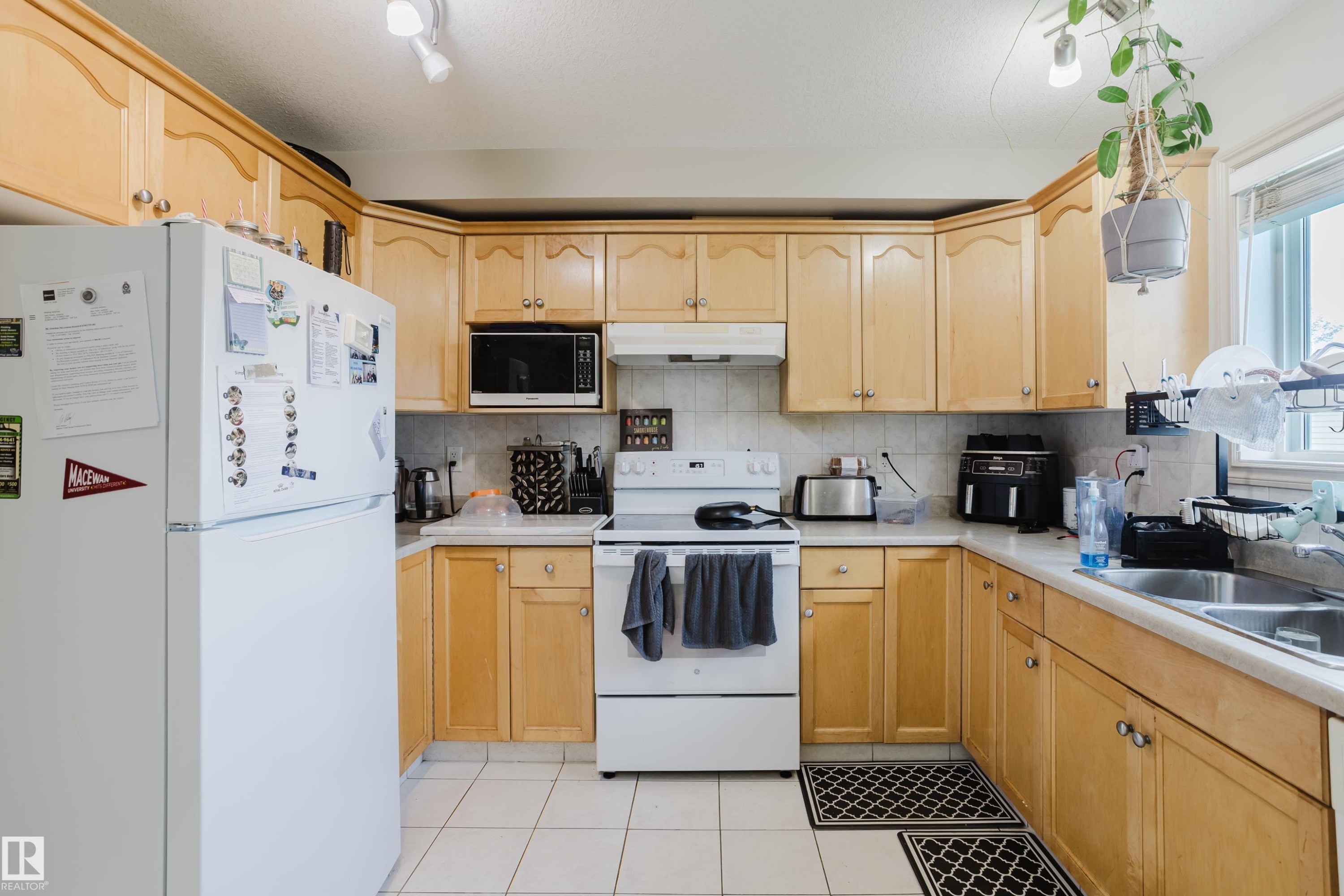U-shaped kitchen featuring wood-finish cabinetry, tile flooring, white appliances, a double basin stainless steel sink, and a tiled backsplash - 10334 161 Street Nw, Edmonton, AB