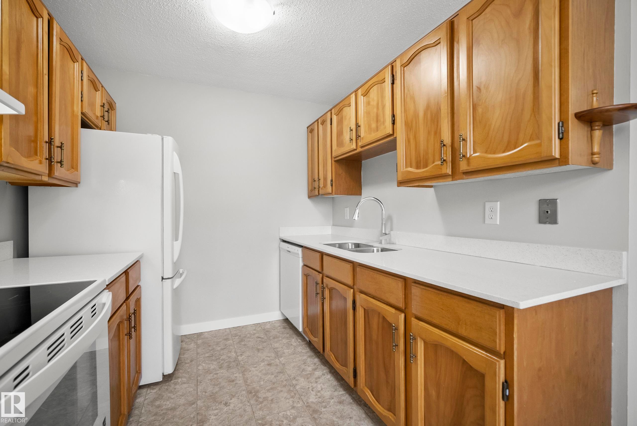 Kitchen featuring wood cabinetry, light-toned countertops, a stainless steel sink, and white appliances - 3173 139 Avenue Nw, Edmonton, AB