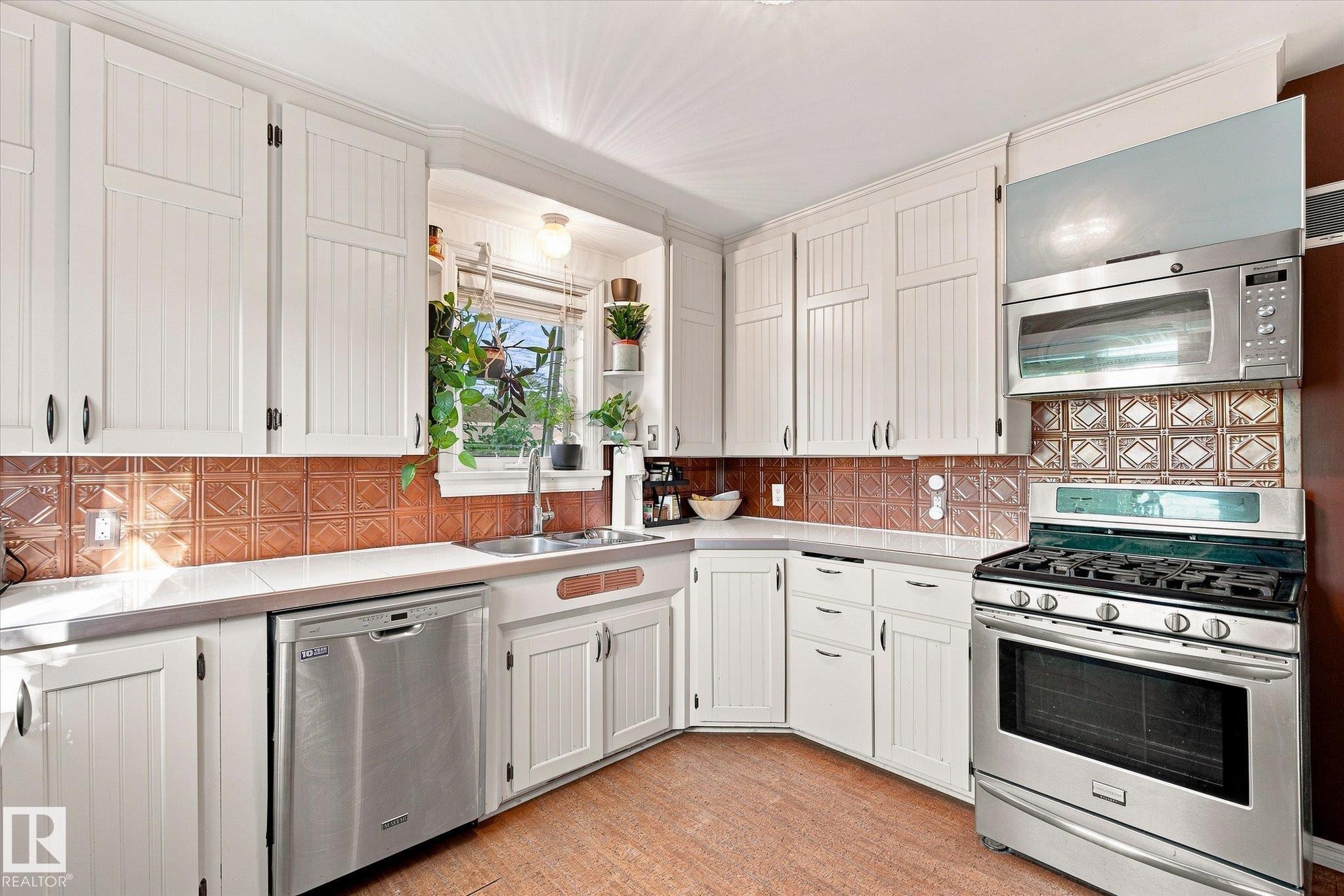 White paneled cabinetry with dark hardware, stainless steel appliances, corner sink with window, copper-toned decorative tile backsplash, and wood-finish flooring - 10943 72 Avenue Nw, Edmonton, AB