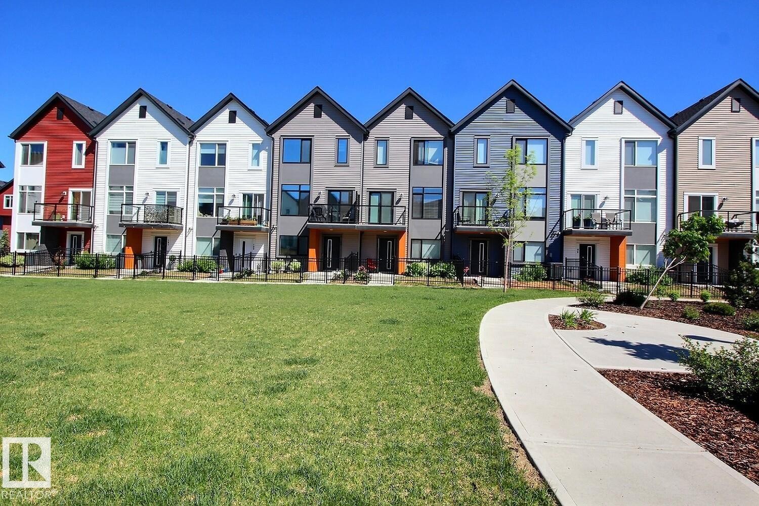 Modern multi-story townhomes featuring contrasting siding, private balconies with glass railings, and a manicured lawn with a concrete pathway - #99 1010 Rabbit Hill Rd Sw, Edmonton, AB