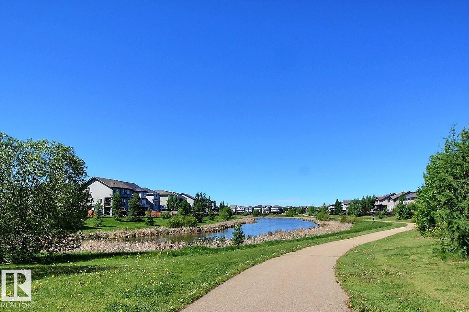 Paved pathway winding alongside a tranquil pond, bordered by green grass and mature trees - #99 1010 Rabbit Hill Rd Sw, Edmonton, AB