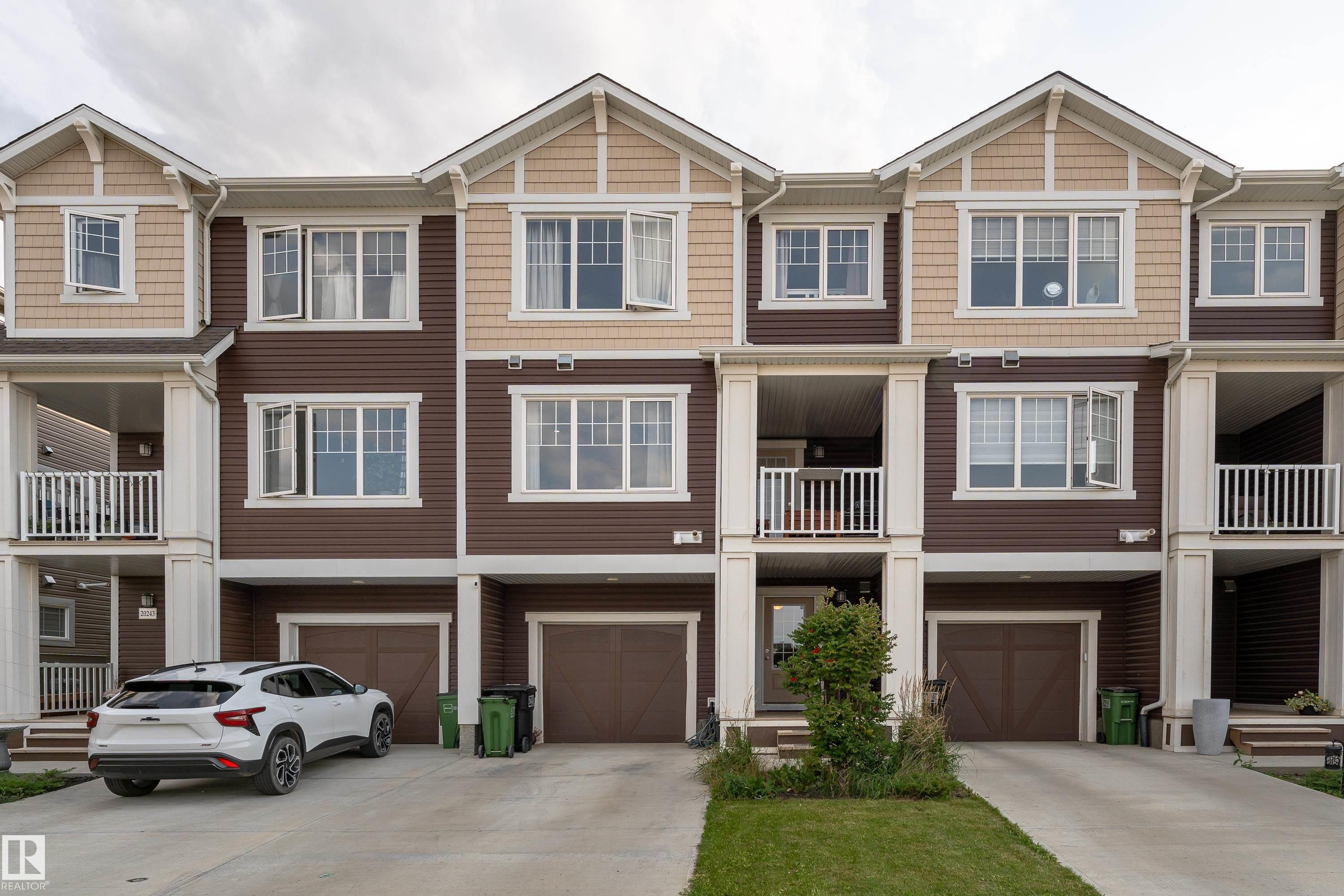 Multi-level townhome featuring a brown and tan exterior, multiple white-framed windows, integrated single-car garages, and covered balconies with white railings - 20247 16 Avenue Nw, Edmonton, AB