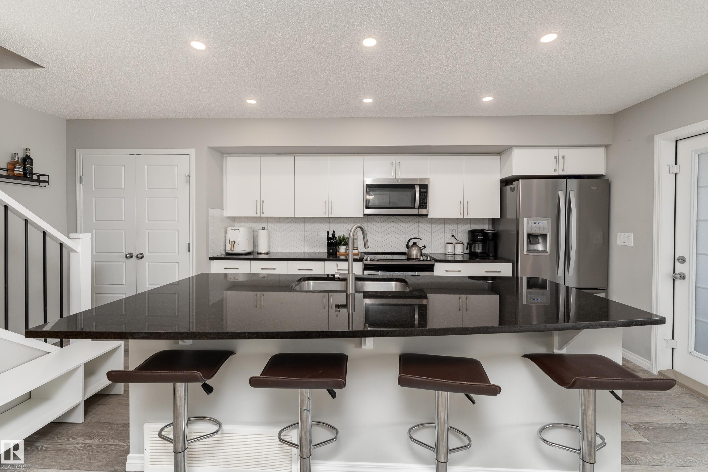Modern kitchen featuring white cabinetry, stainless steel appliances, a dark countertop island, a herringbone tile backsplash, and recessed lighting - 20247 16 Avenue Nw, Edmonton, AB