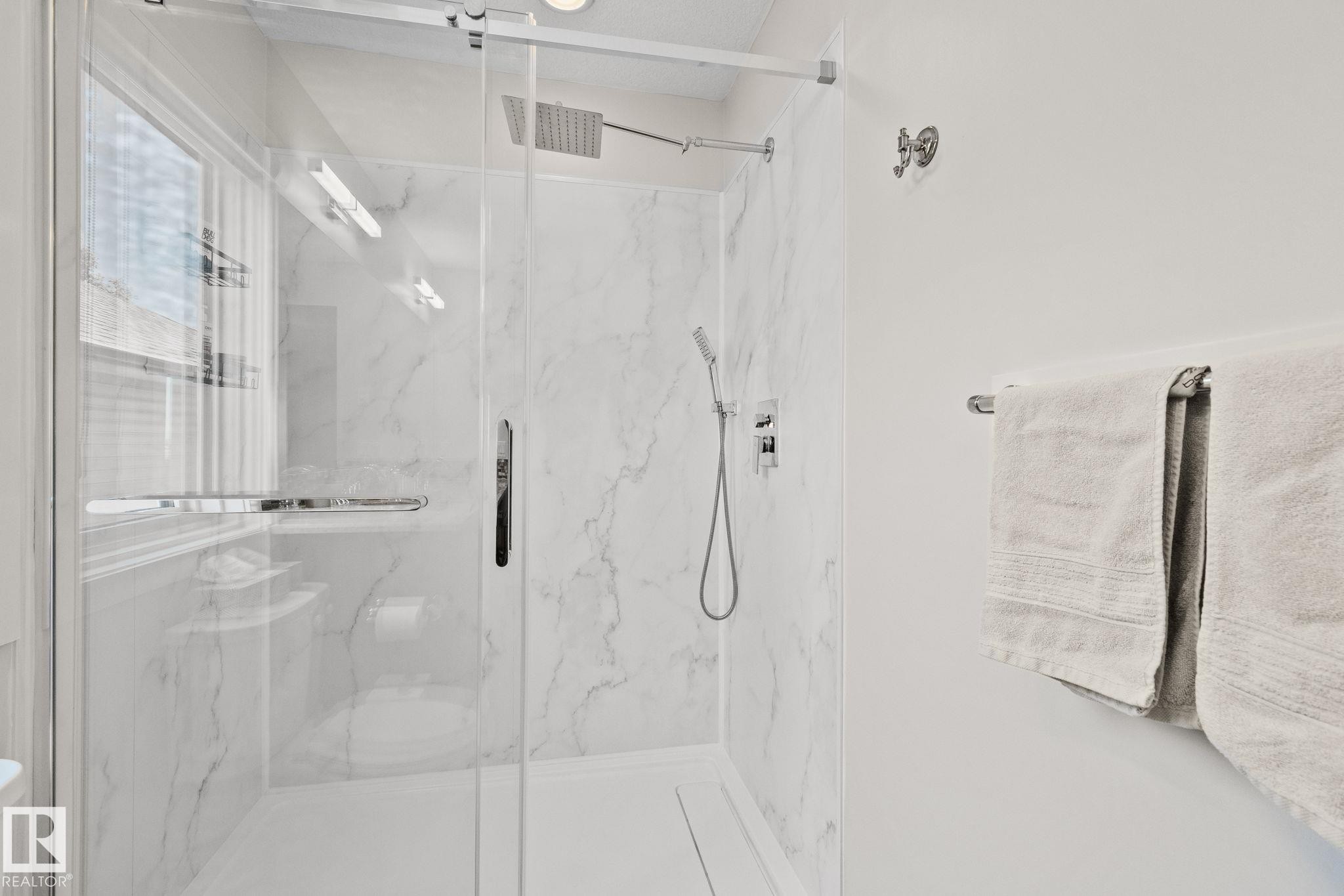 Walk-in shower with clear glass enclosure, marble-finish wall panels, and chrome fixtures including a rain showerhead and handheld sprayer - 2096 Redtail Common Nw, Edmonton, AB