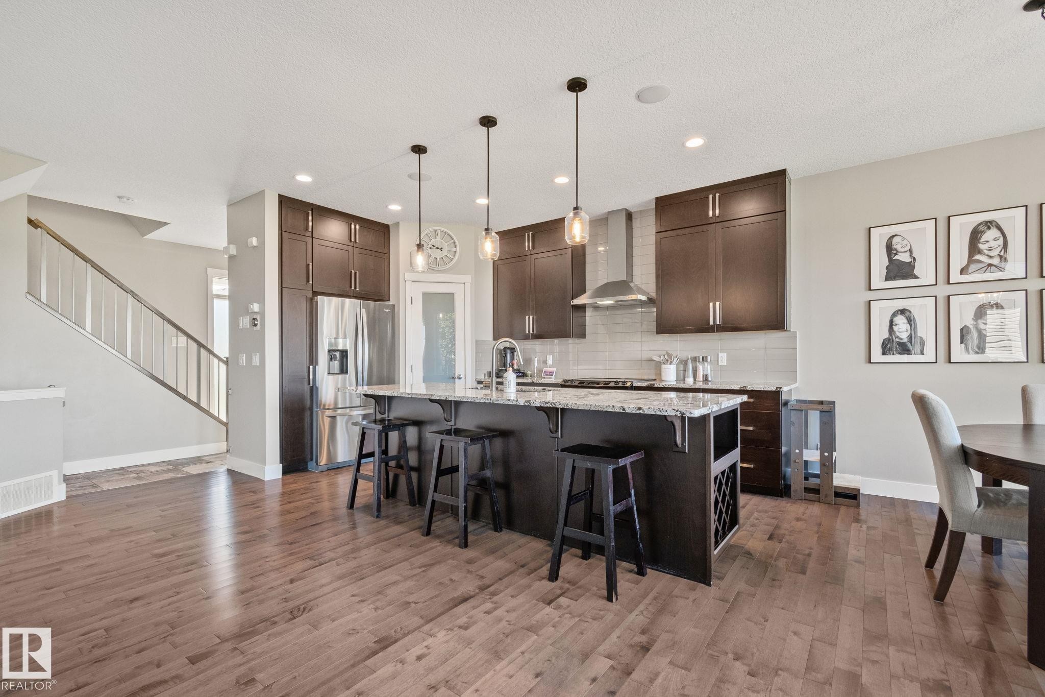 Kitchen with dark wood-finish cabinetry, stainless steel appliances, a multi-person island with a stone countertop, and wood-finish flooring - 2096 Redtail Common Nw, Edmonton, AB