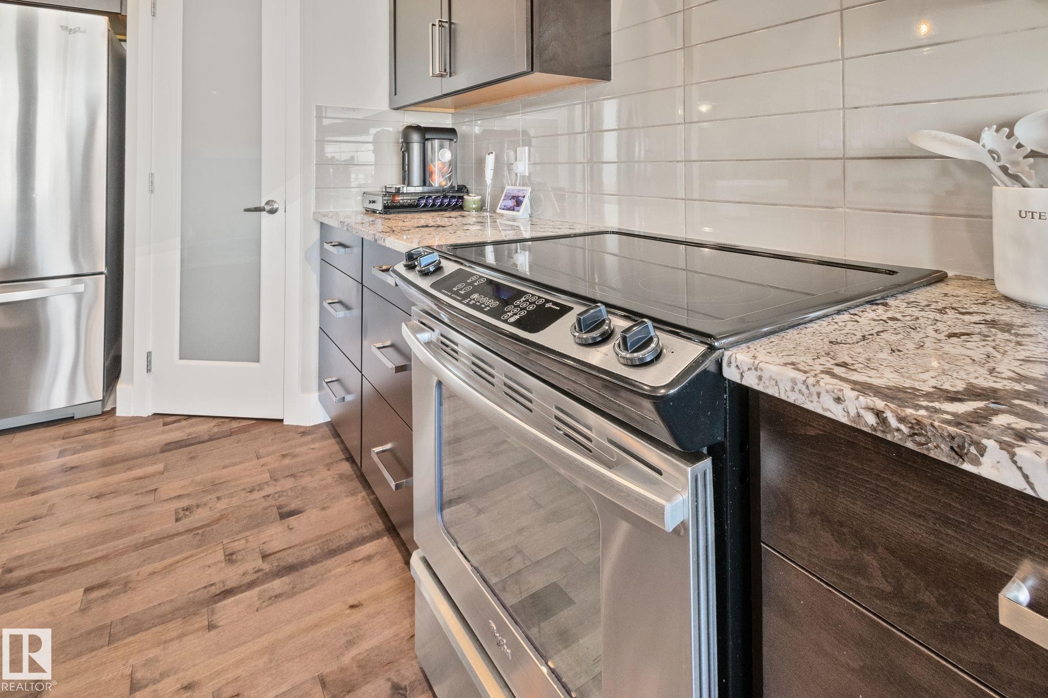 Stainless steel range with a smooth cooktop, wood-finish flooring, dark wood cabinetry, light-toned stone countertops, and a white subway tile backsplash - 2096 Redtail Common Nw, Edmonton, AB