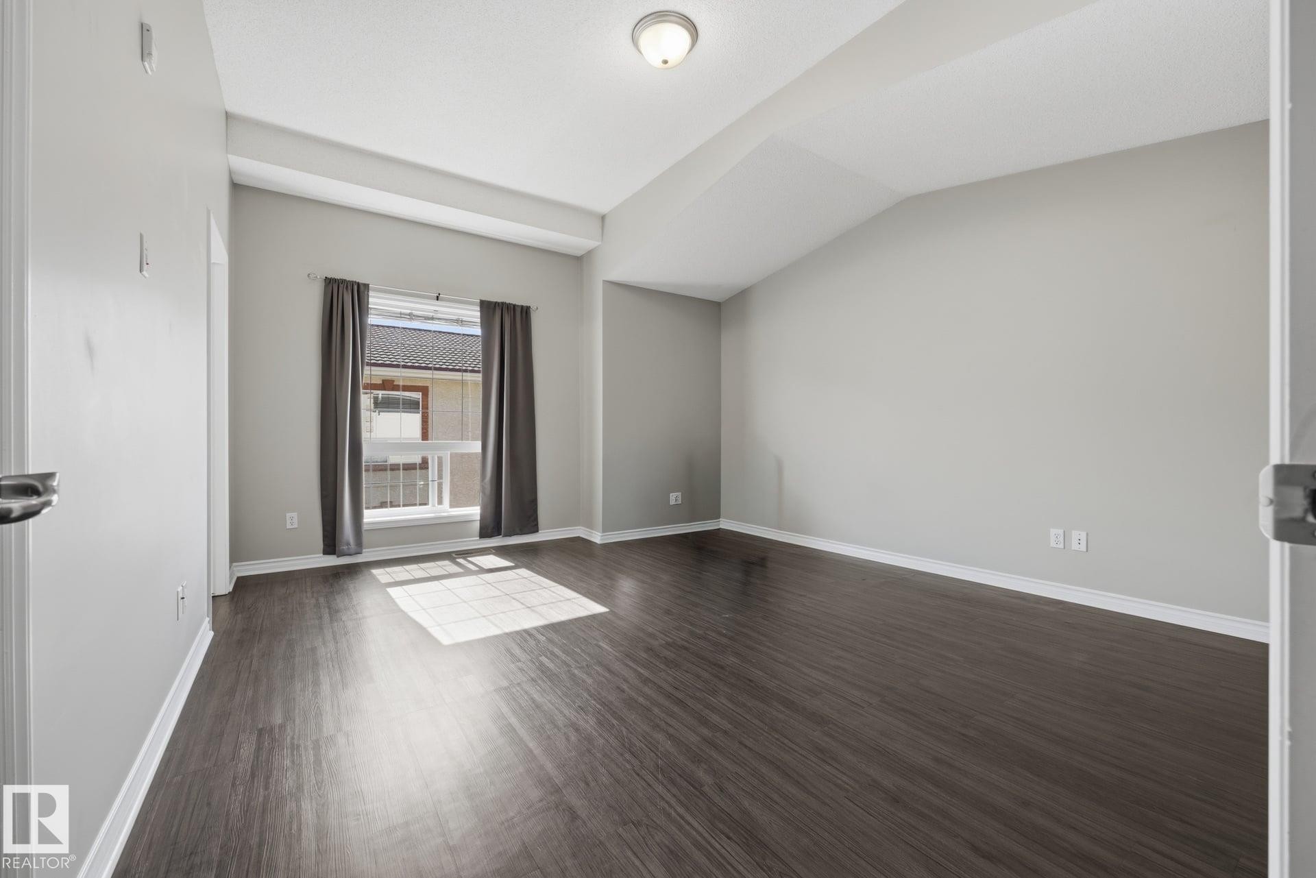 Vaulted ceiling with an exposed beam, wood-finish flooring, neutral wall paint, and a window with curtain rods - #102 13825 155 Av Nw, Edmonton, AB