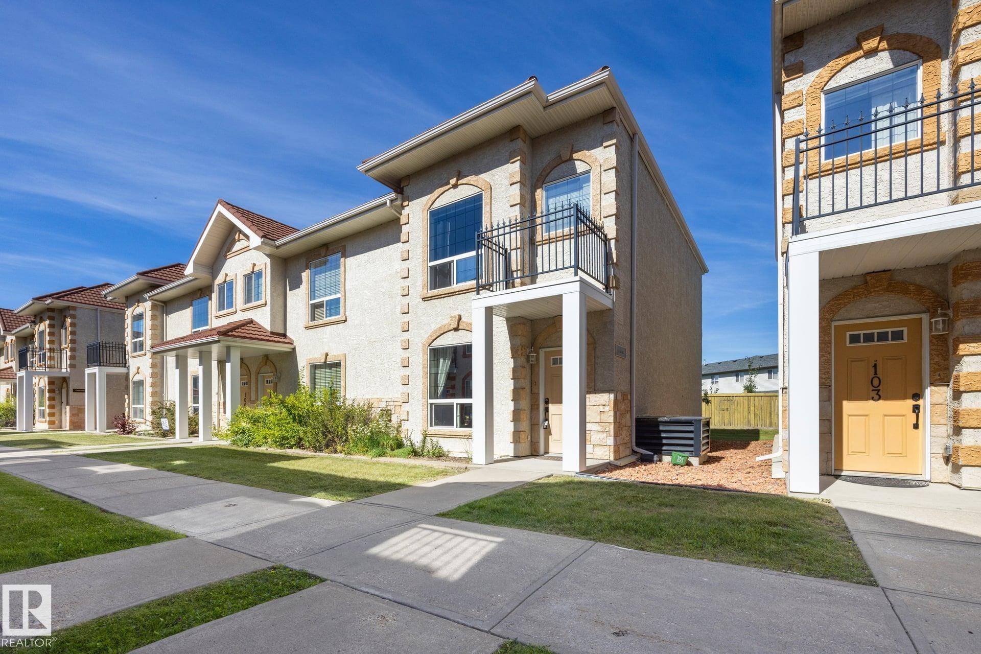 Stucco exterior with stone accents, arched windows, second-story balcony with metal railing, and covered entry - #102 13825 155 Av Nw, Edmonton, AB