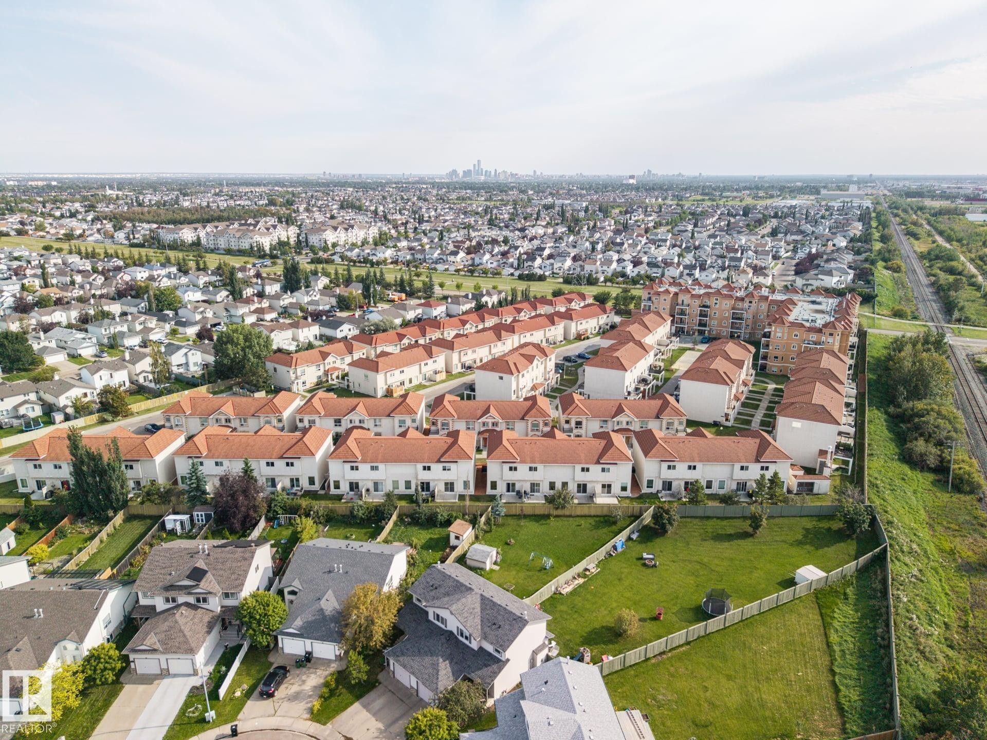 Aerial perspective showcasing a residential community with red-tiled roofs, a large grass area, and a distant city skyline - #102 13825 155 Av Nw, Edmonton, AB