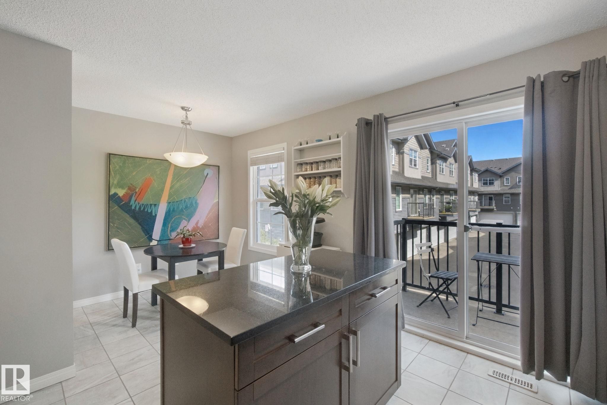 Kitchen island with dark countertop, dining area with pendant lighting, tile flooring, built-in shelving, and sliding glass door with balcony - #58 4029 Orchards Dr Sw, Edmonton, AB