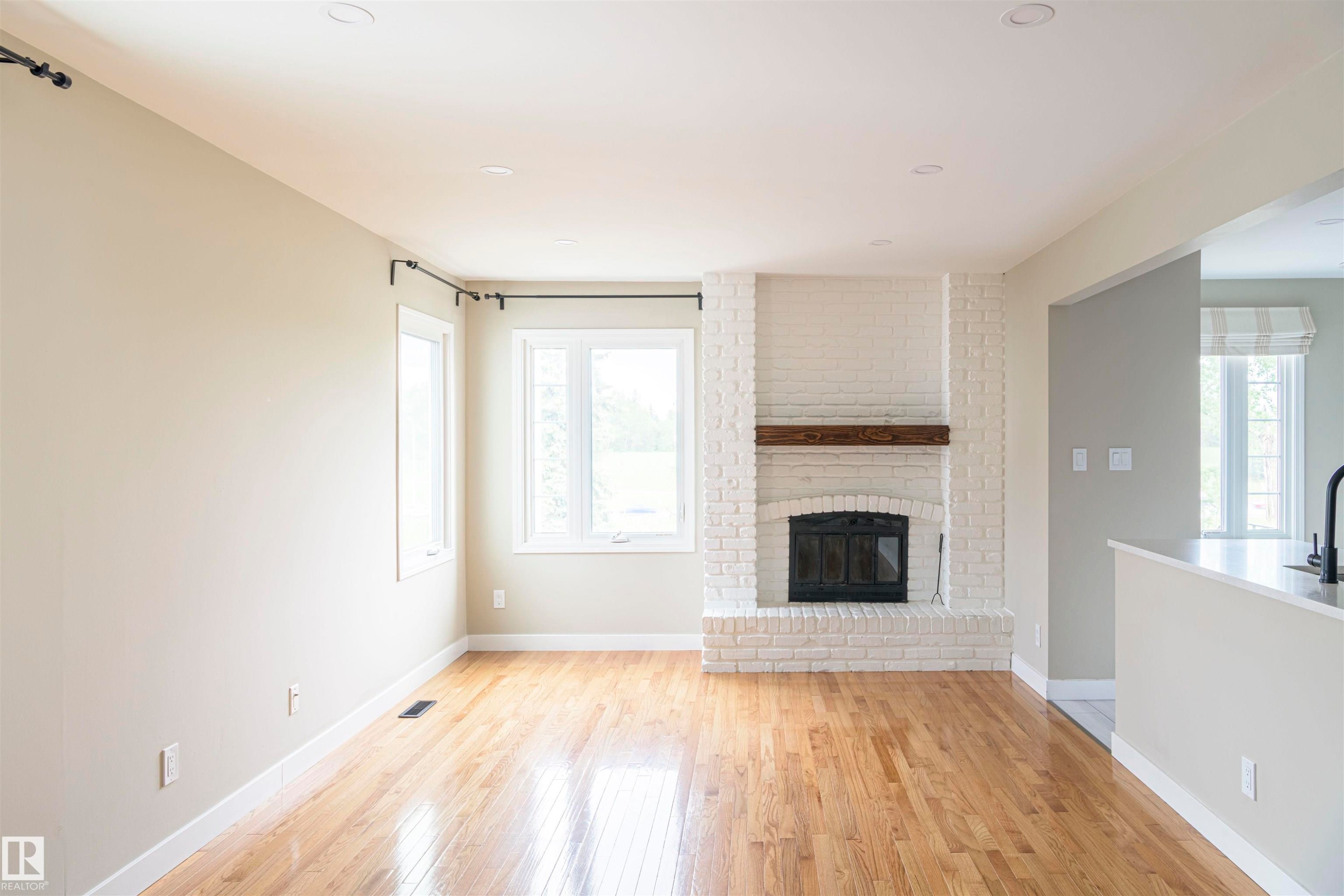 Spacious room featuring wood-finish flooring, a white-painted brick fireplace with a wood mantel, recessed lighting, and multiple windows - 5010 147 Street Nw, Edmonton, AB