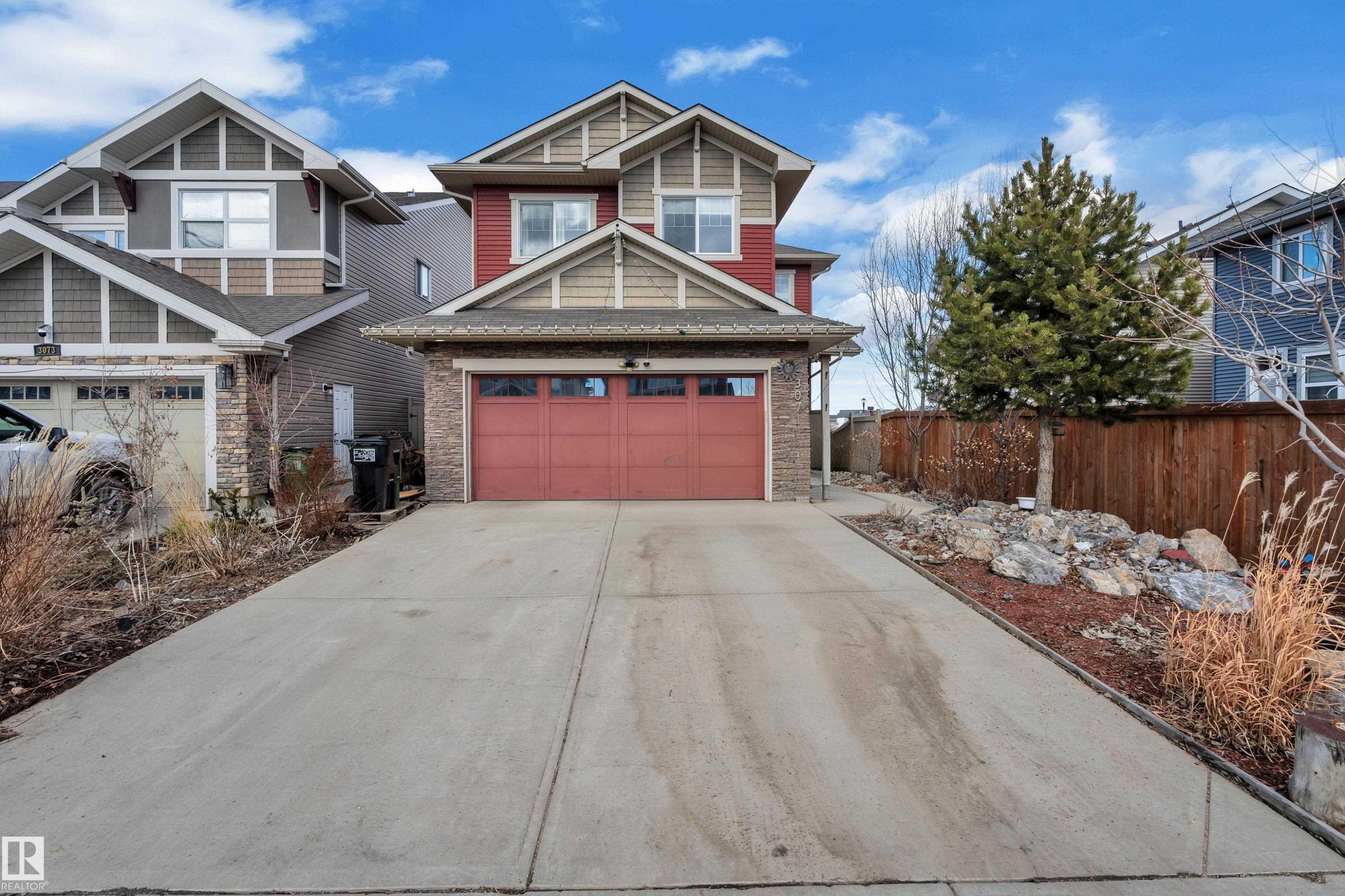 Two-story exterior with red siding and light brown shake accents, featuring a two-car garage with a red door, a concrete driveway, and a rock garden with mature evergreen landscaping - 3071 Carpenter Landing Sw, Edmonton, AB