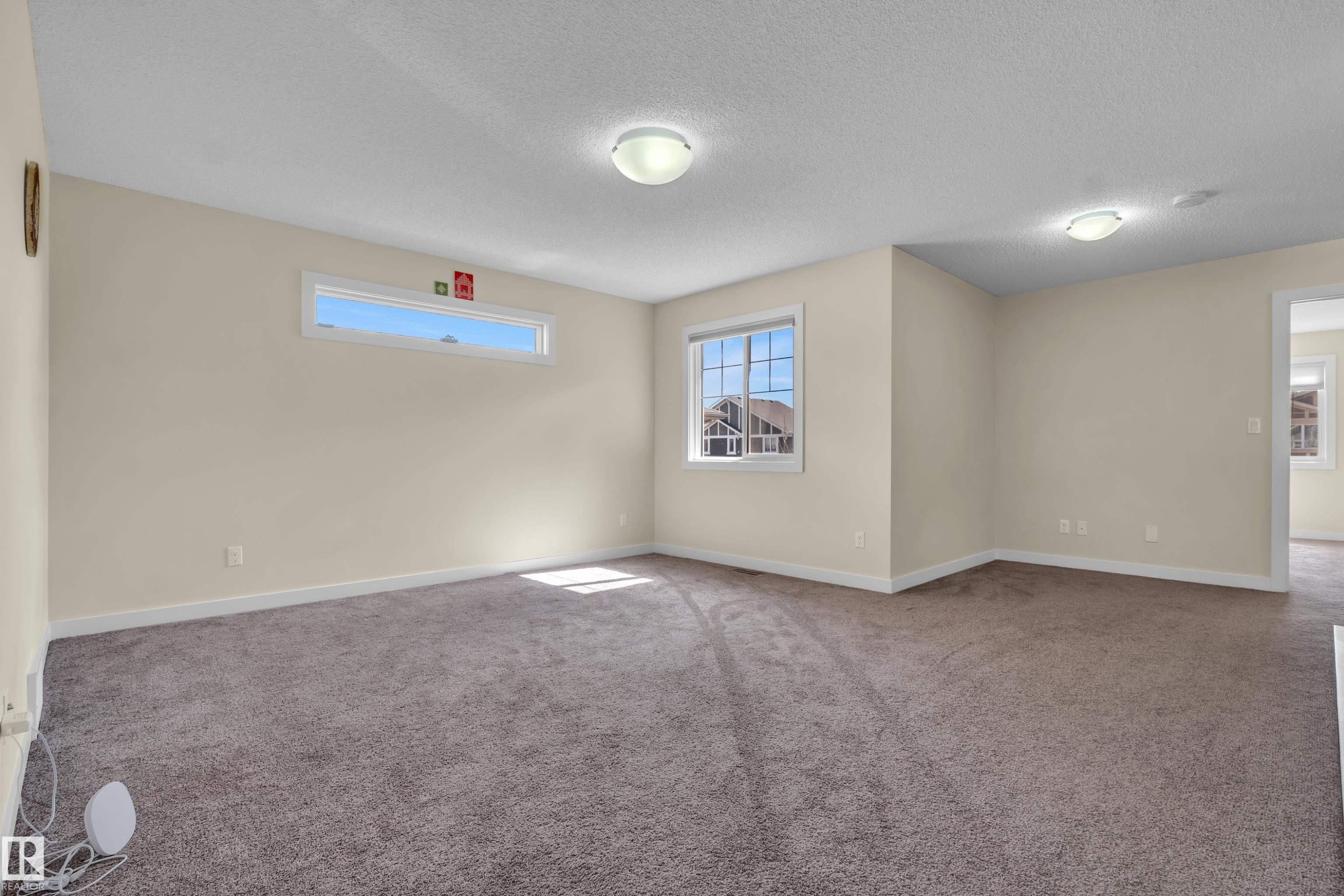Spacious room featuring neutral carpeting, light beige walls, multiple windows, and flush-mount ceiling lighting - 3071 Carpenter Landing Sw, Edmonton, AB
