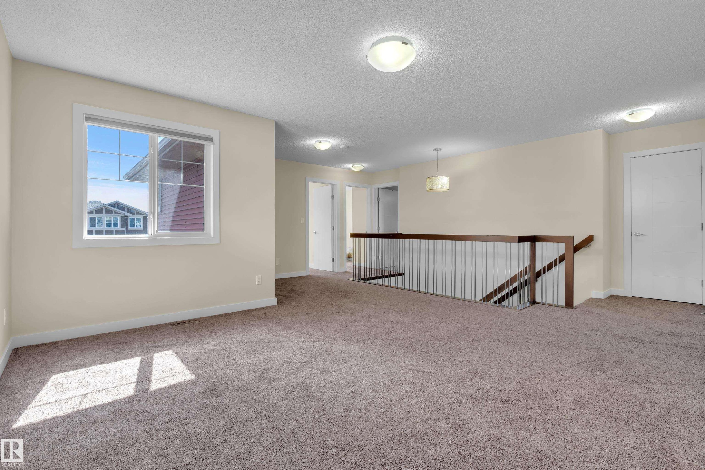 Spacious carpeted landing area featuring a large window, white trim, a wood and metal railing, and multiple flush-mount ceiling lights - 3071 Carpenter Landing Sw, Edmonton, AB