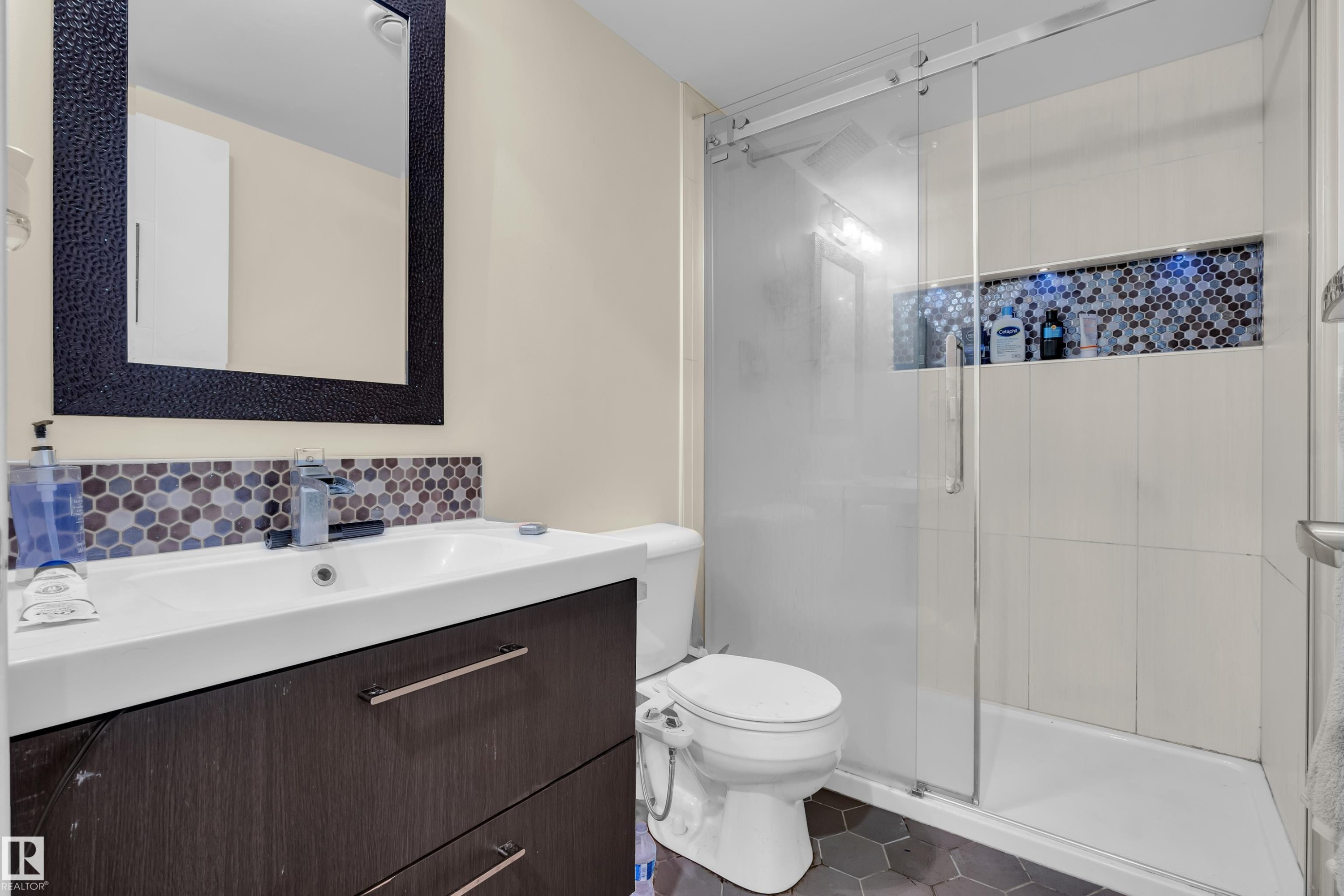 Contemporary bathroom featuring a dark wood-finish vanity with an integrated white sink, a mosaic tile backsplash, a frameless mirror with a textured black frame, a glass-enclosed shower with subway tile surround, and hexagonal floor tiles - 3071 Carpenter Landing Sw, Edmonton, AB