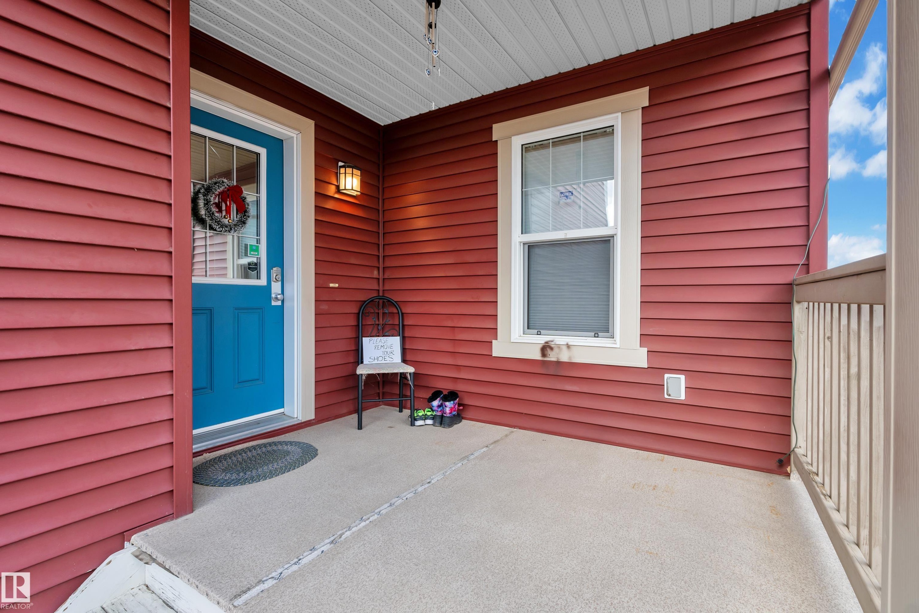 Inviting front porch featuring a vibrant blue entry door with decorative glass, red horizontal siding, a white-framed window, a built-in outdoor light fixture, and a light-colored porch railing - 3071 Carpenter Landing Sw, Edmonton, AB