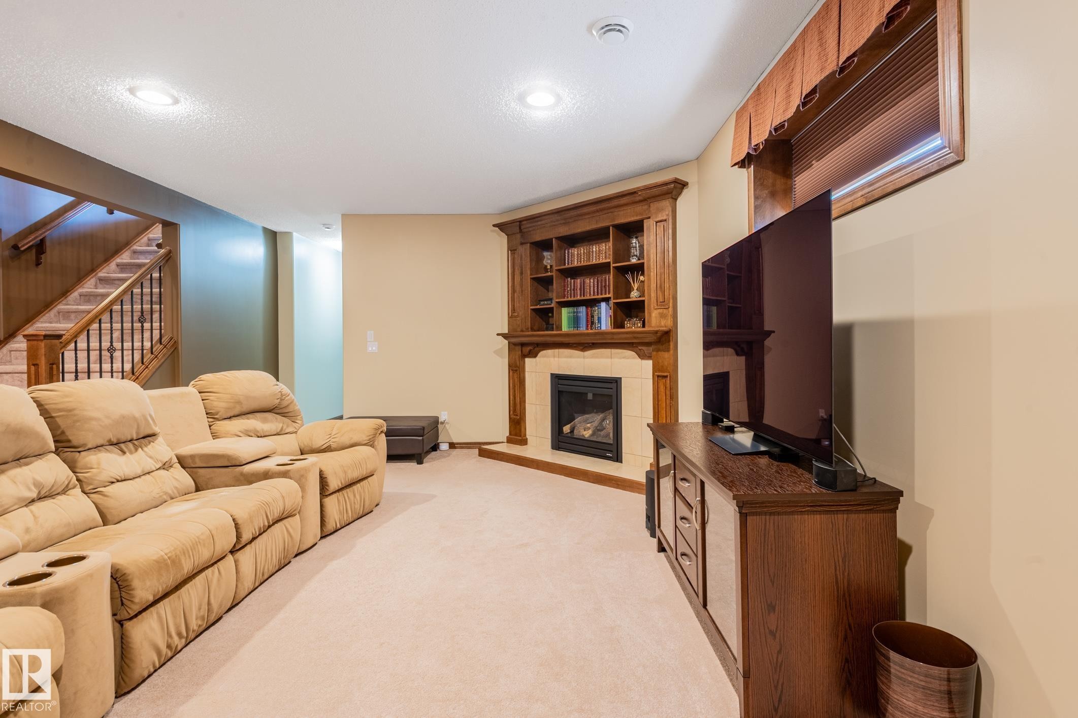 Carpeted recreation room featuring a corner fireplace with a tile surround and wood mantel, built-in shelving, a wood-finish media console, and a staircase with wood newel posts and handrails - 19 Newton Place, St. Albert, AB