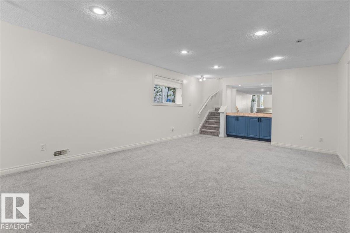 Spacious room with recessed lighting, plush carpeting, a built-in wet bar with wood-finish countertop and blue cabinetry, and a carpeted staircase with a white railing - 15420 44 Street Nw, Edmonton, AB