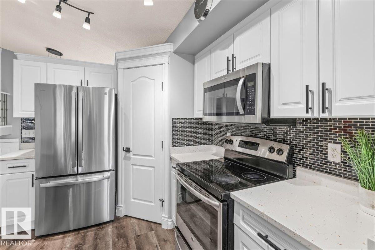 White cabinetry with black hardware, stainless steel appliances, dark mosaic tile backsplash, light-toned countertops, and wood-finish flooring - 15420 44 Street Nw, Edmonton, AB
