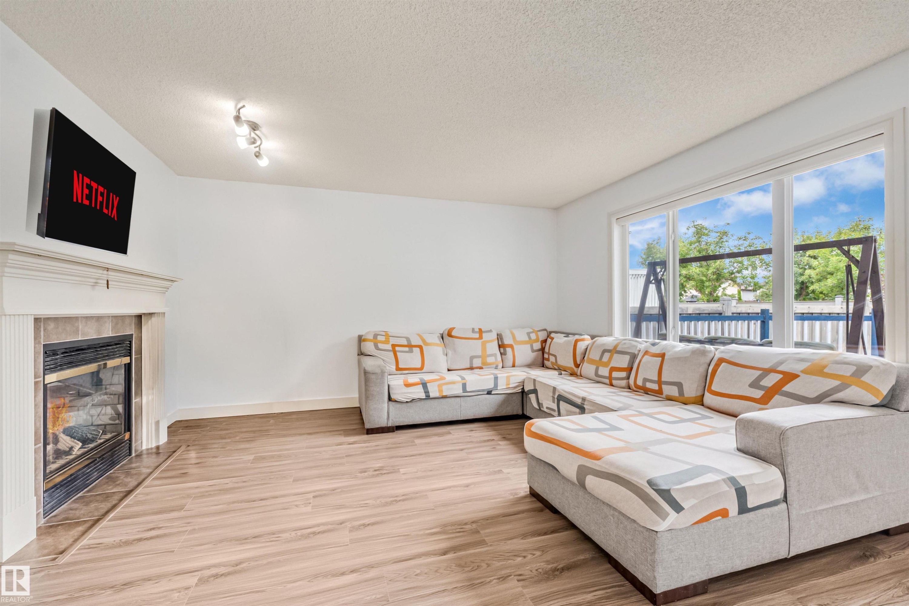 Living area with wood-finish flooring, a tiled fireplace with a white mantel, and large picture windows - 20324 48 Avenue Nw, Edmonton, AB
