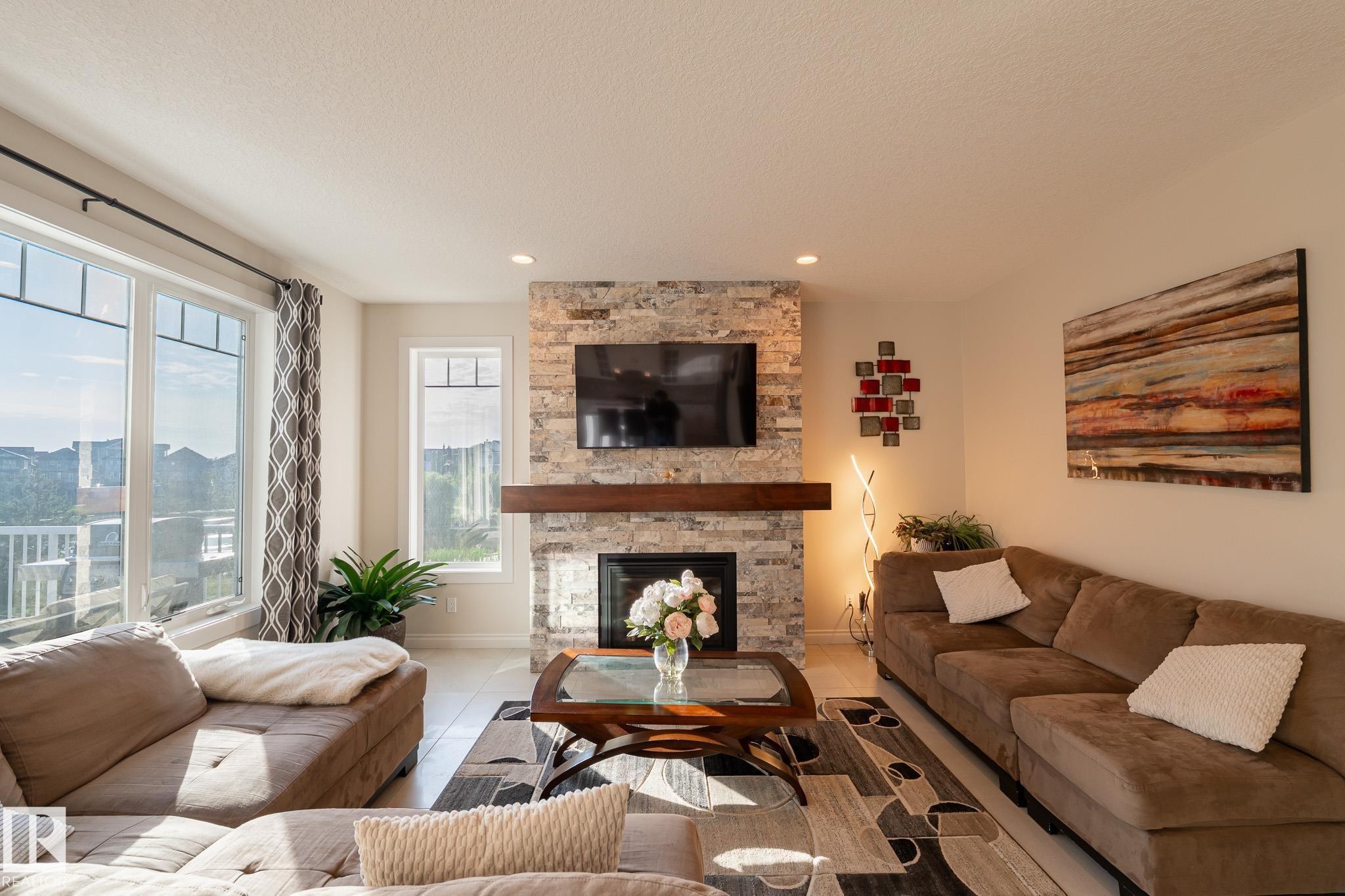 Spacious living area featuring a stone-clad fireplace with a wood mantel, built-in recessed lighting, large windows, and light-toned flooring - 914 Crystallina Nera Way Nw, Edmonton, AB