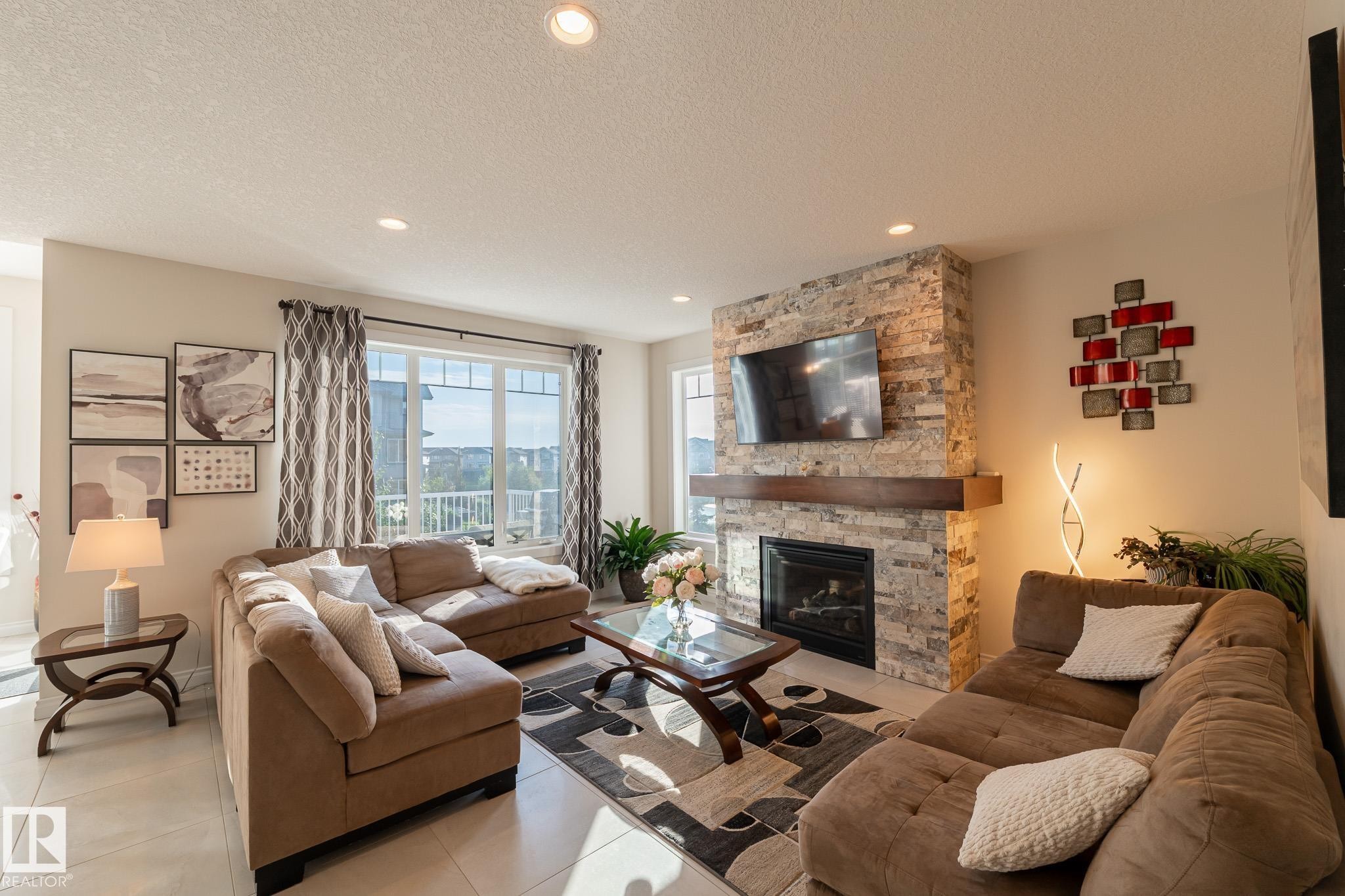 Living area featuring a stone-clad fireplace with a wood mantel, recessed lighting, and large windows with horizontal mullions - 914 Crystallina Nera Way Nw, Edmonton, AB