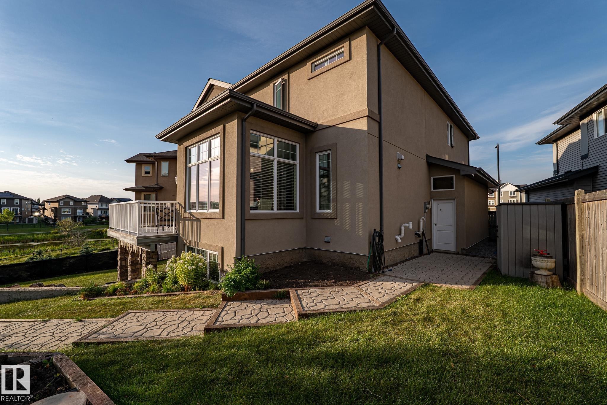 Stucco exterior with multiple windows, elevated deck with white railing, patterned paver patio, and sloped grassy area - 914 Crystallina Nera Way Nw, Edmonton, AB