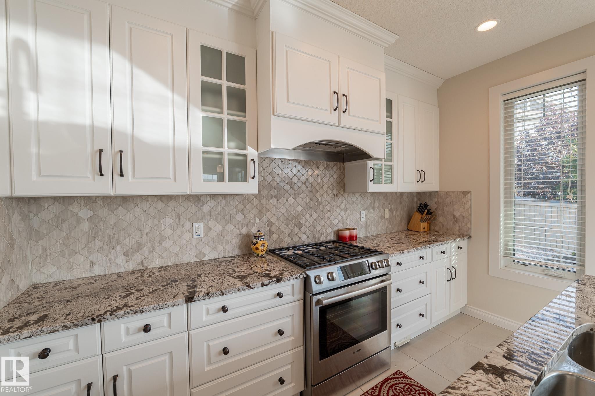 White cabinetry with dark hardware, stone countertops, geometric tile backsplash, stainless steel range, and an undermount sink - 914 Crystallina Nera Way Nw, Edmonton, AB
