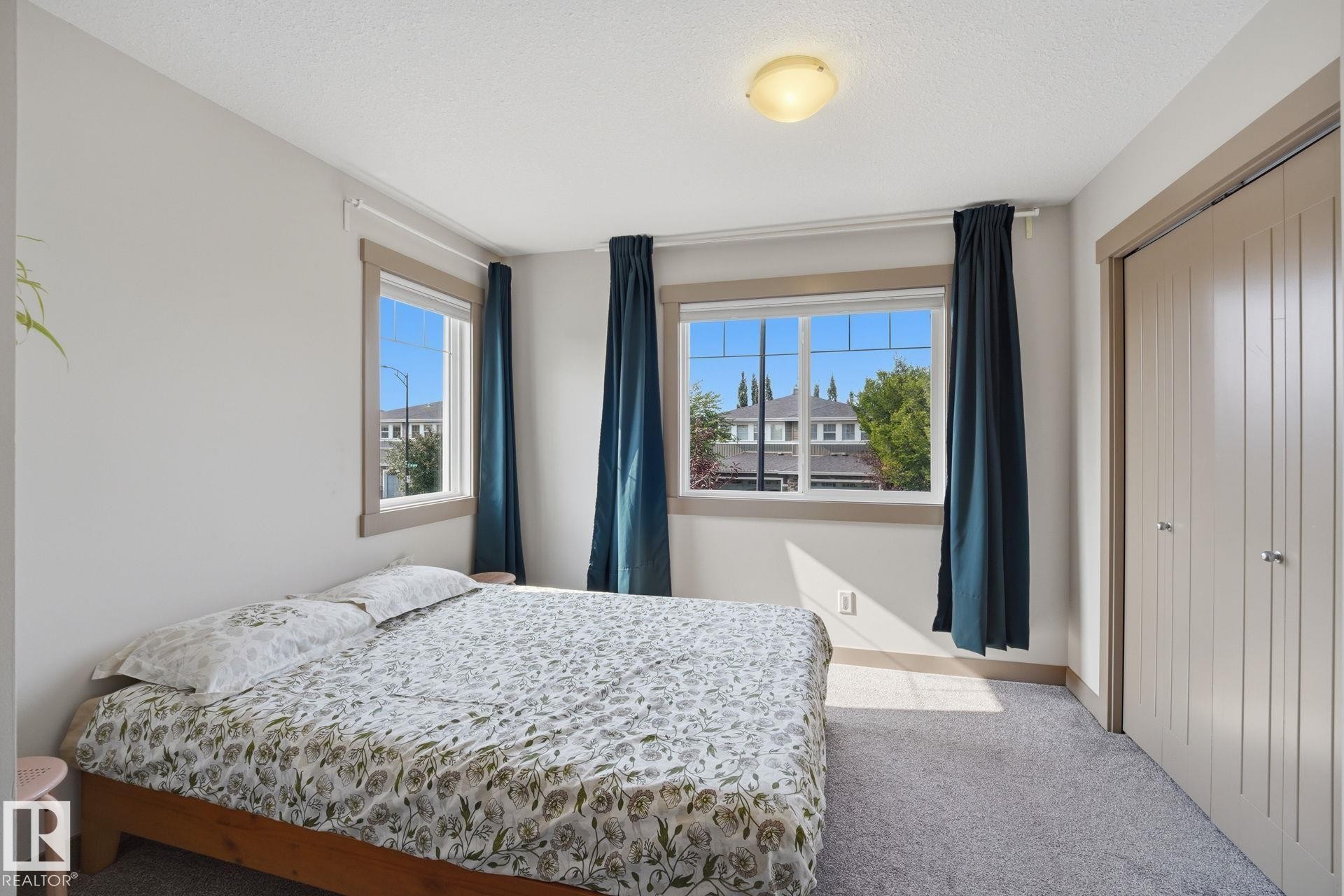Carpeted bedroom featuring multiple windows with wood-finish trim, a flush-mount ceiling light, and a built-in closet with vertical panel doors - 17621 10 Avenue Sw, Edmonton, AB - Indoor Photo Showing Bedroom