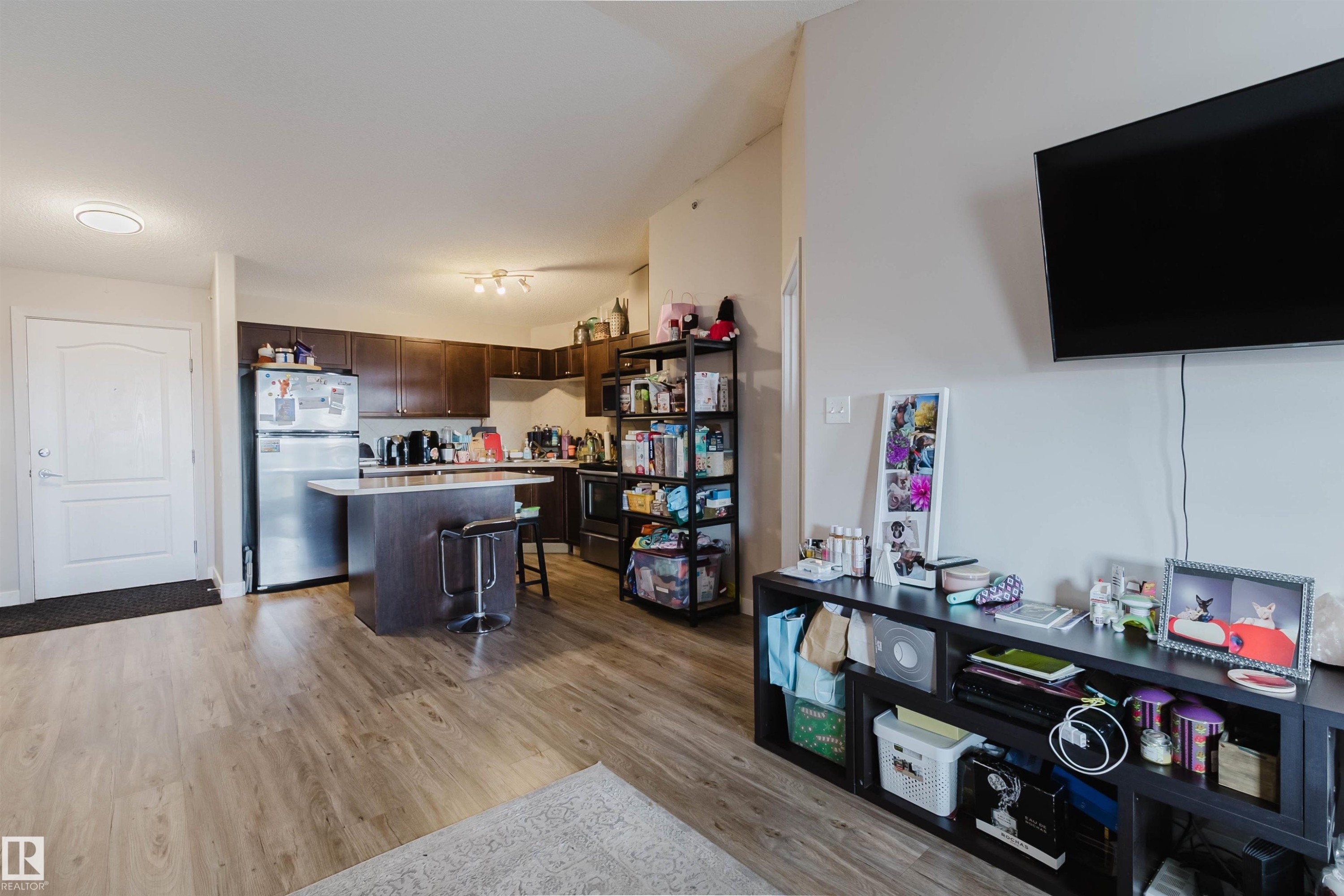 Open-concept living area with wood-finish flooring, a kitchen island with seating, stainless steel refrigerator, and dark wood cabinetry - #7428 7327 South Terwillegar Dr Nw, Edmonton, AB