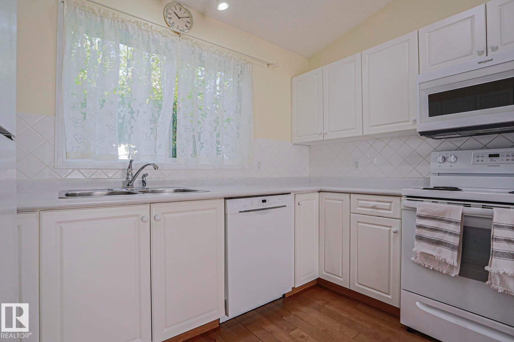 Kitchen featuring white cabinetry, a built-in microwave, a double basin sink, white appliances, and wood-finish flooring - 15031 135 Street Nw, Edmonton, AB