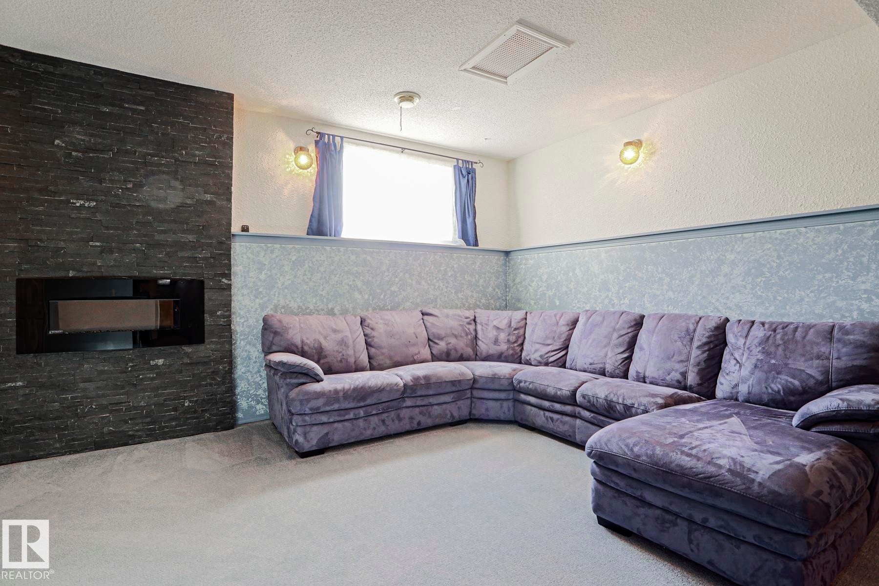 Stacked stone-finish fireplace with an integrated firebox and hearth, wall-mounted light fixtures, textured wall covering, and a window for natural light - 15031 135 Street Nw, Edmonton, AB