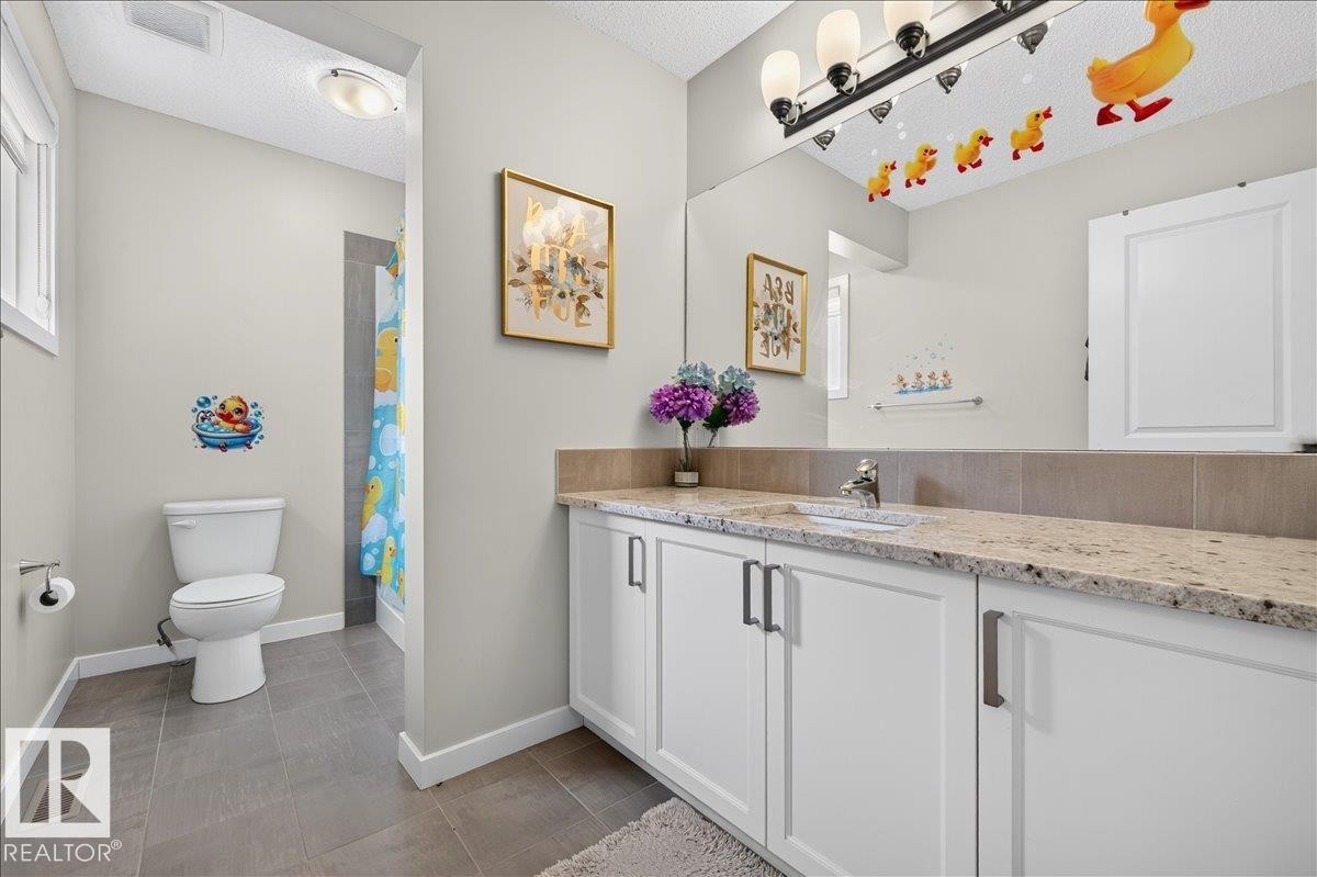 Bathroom featuring a vanity with a light-toned, speckled countertop, white cabinetry with dark hardware, a large mirror, a white toilet, and gray tile flooring - 2709 Collins Crescent Sw, Edmonton, AB - Indoor Photo Showing Bathroom