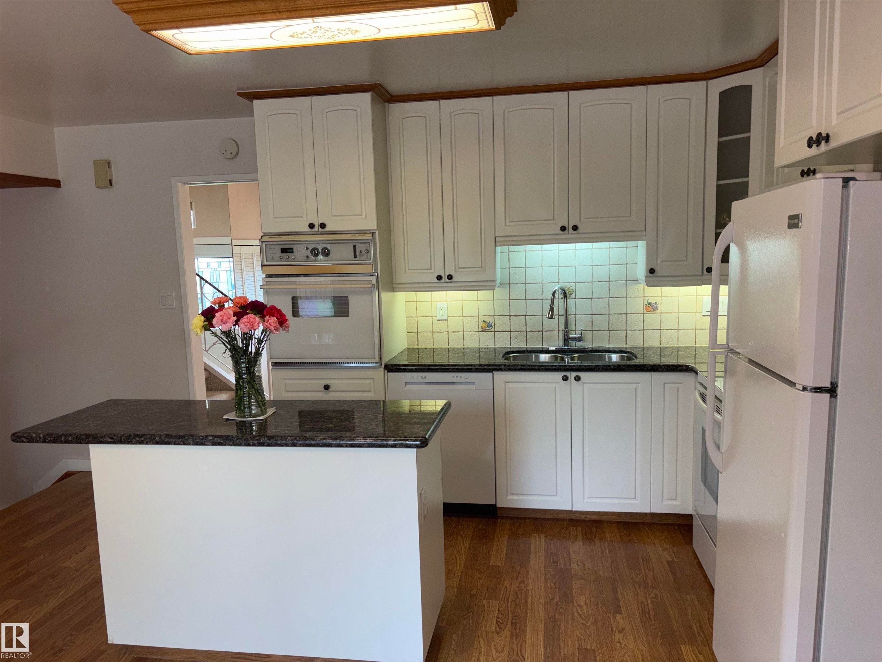 Kitchen featuring white cabinetry with dark hardware, granite-look countertops, a built-in oven, and wood-finish flooring - 4027 74 Street Nw, Edmonton, AB